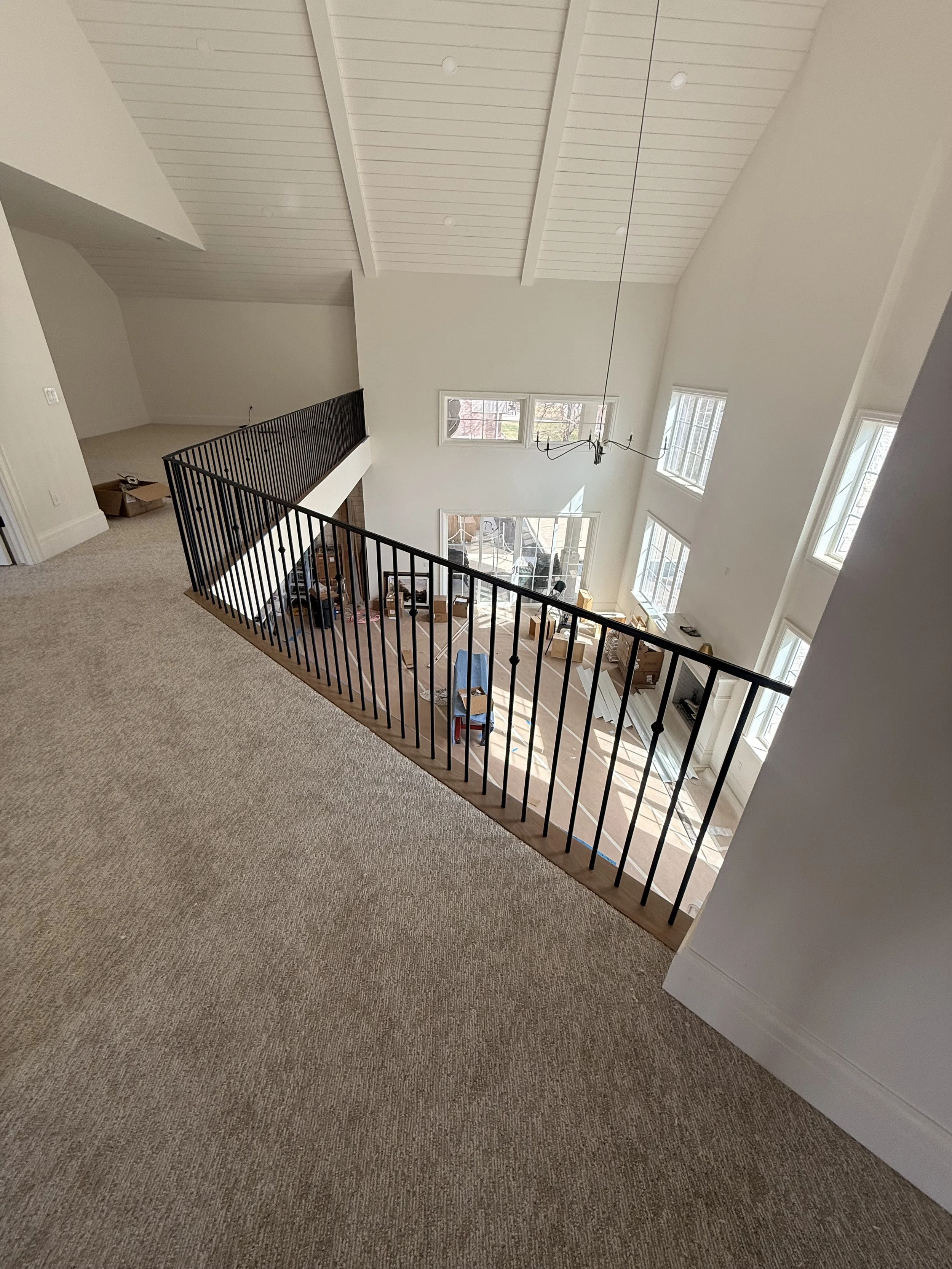 Interior view of a multi-story house with a beige carpeted floor on the upper level, black metal railing separating the upper and lower levels, and large windows allowing natural light into the living space below. The ceiling has white wooden panels 