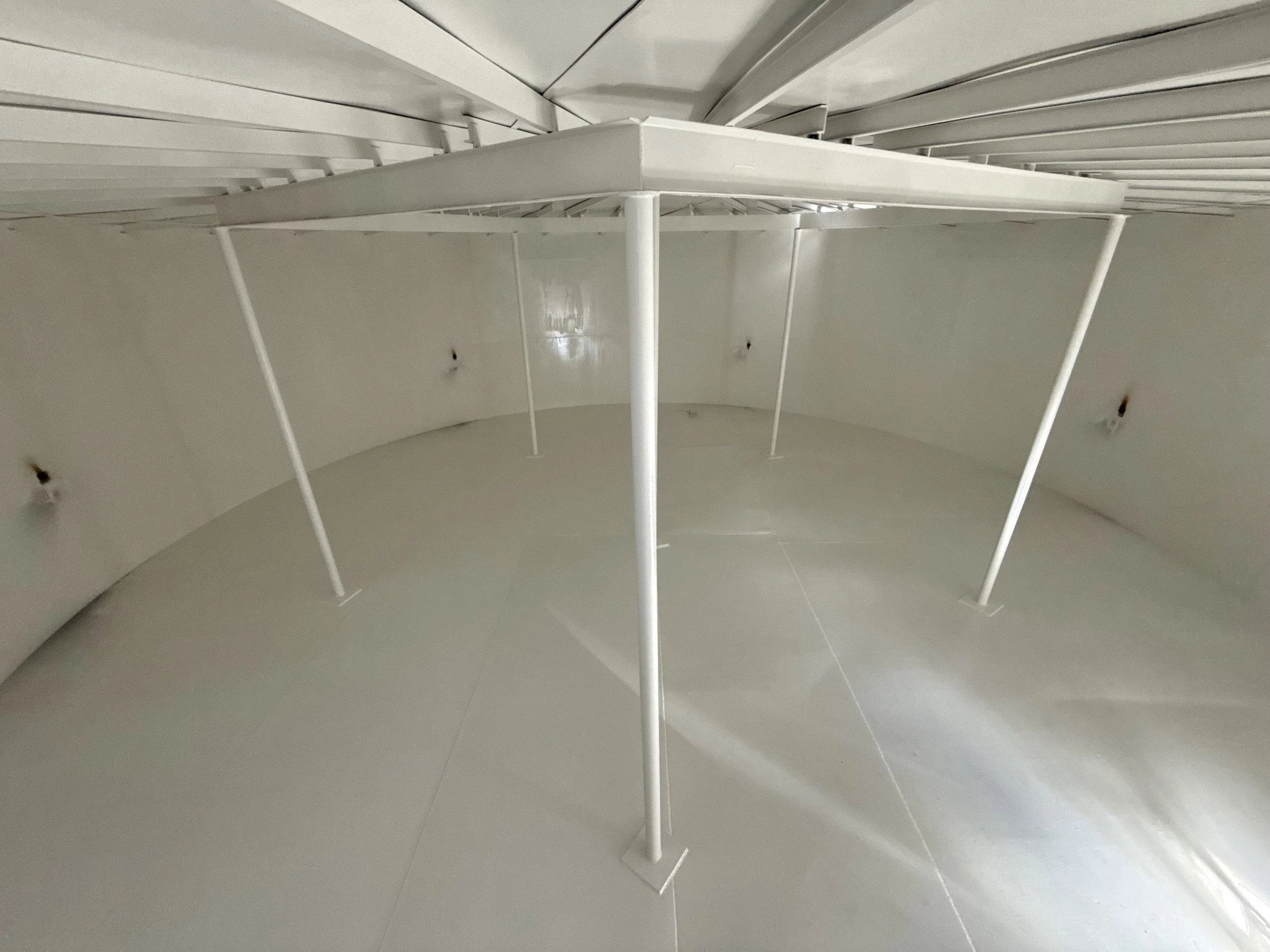Interior of an empty, white, cylindrical industrial storage tank or silo with metal support beams and a smooth floor.