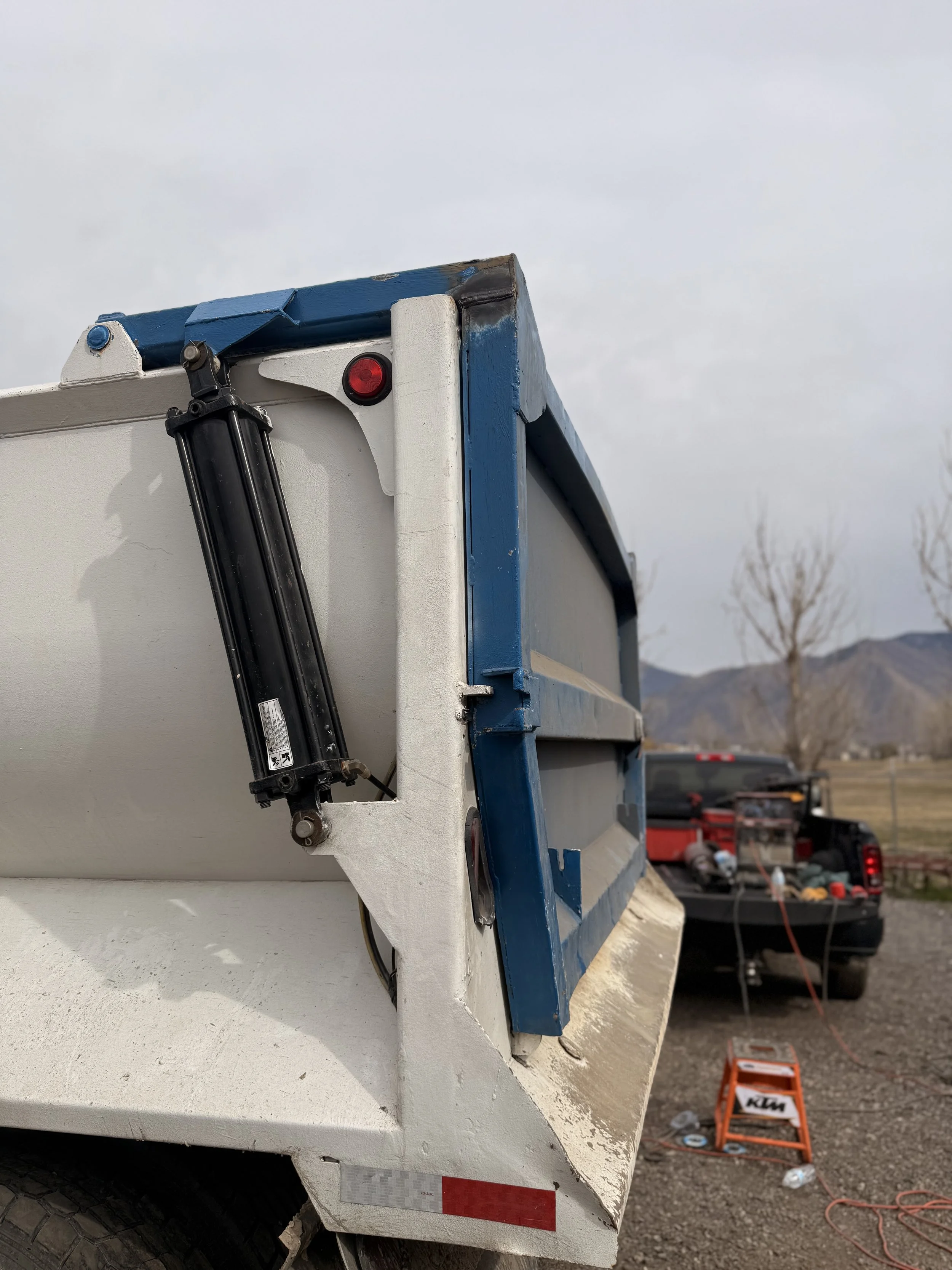 Close-up of the rear corner of a truck bed showing a blue and white structure with a hydraulic lift mechanism, and a black truck in the background in a rural area with mountains and leafless trees.