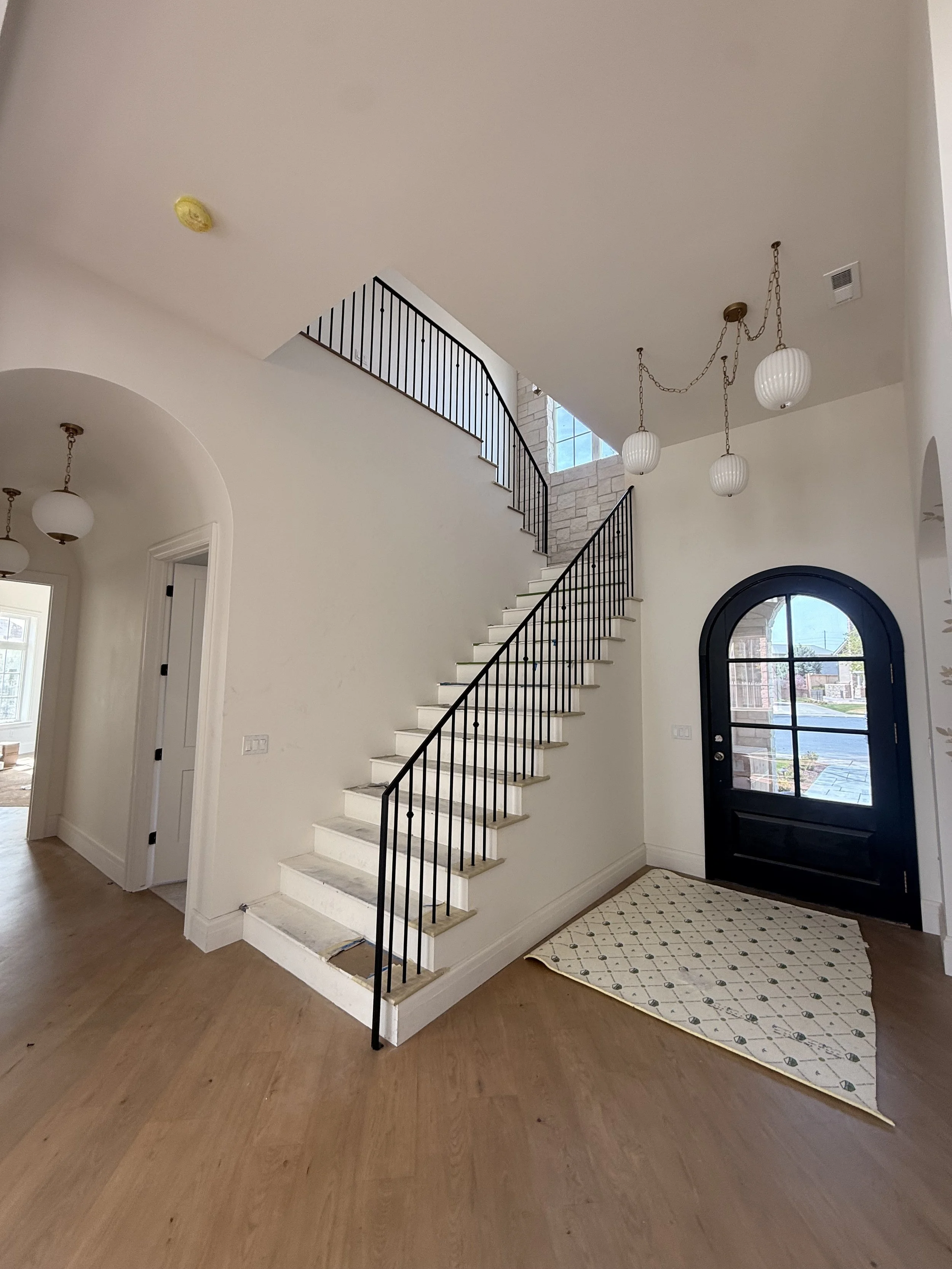Interior view of a modern entryway with a staircase, black front door with window, hardwood flooring, and a patterned rug. There are pendant lights hanging from the ceiling and a fire alarm on the ceiling.