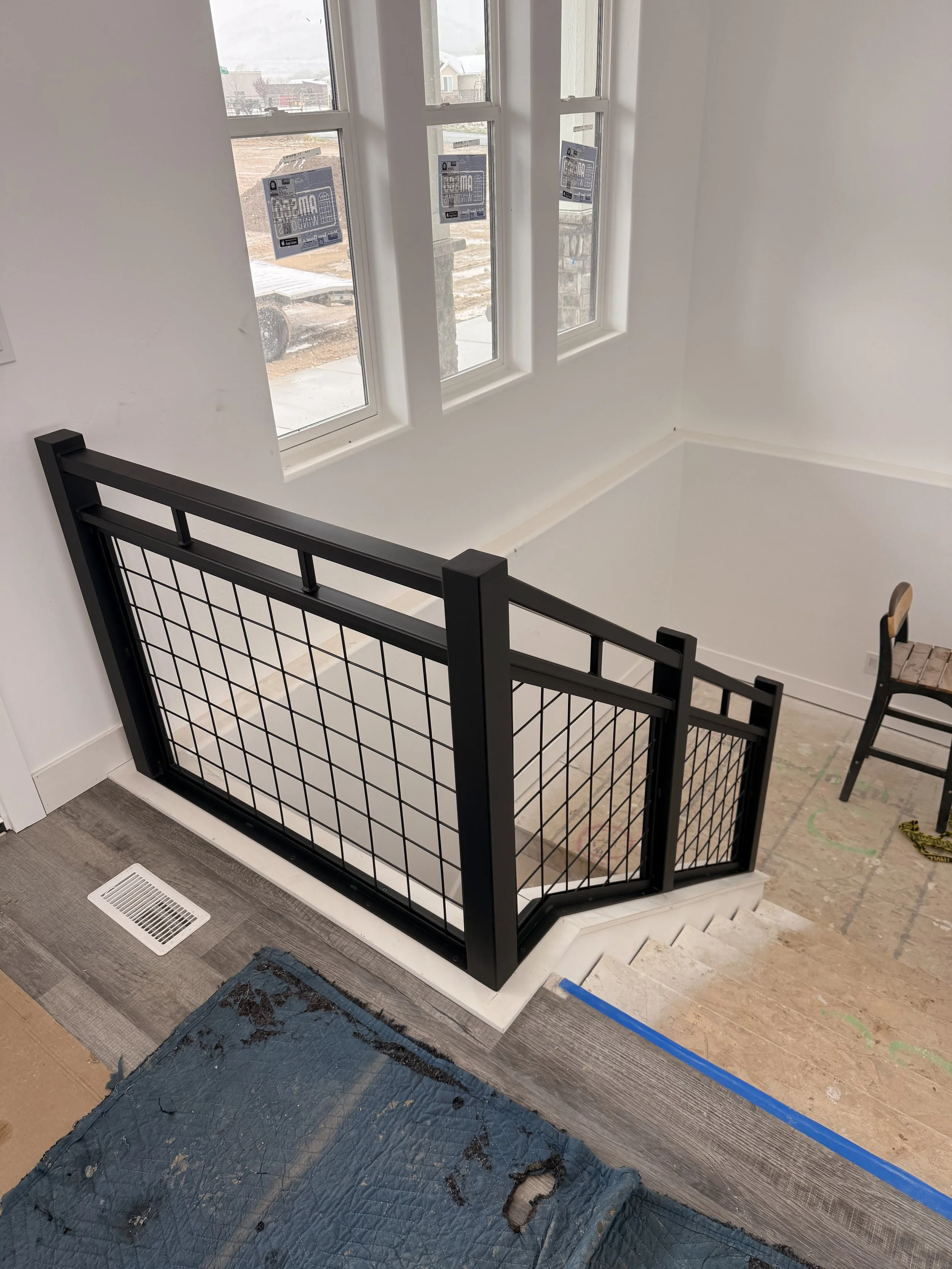 Interior view of a staircase with black metal railing, white walls, and large windows overlooking a construction site outside. There is a wooden chair and some construction materials on the floor.