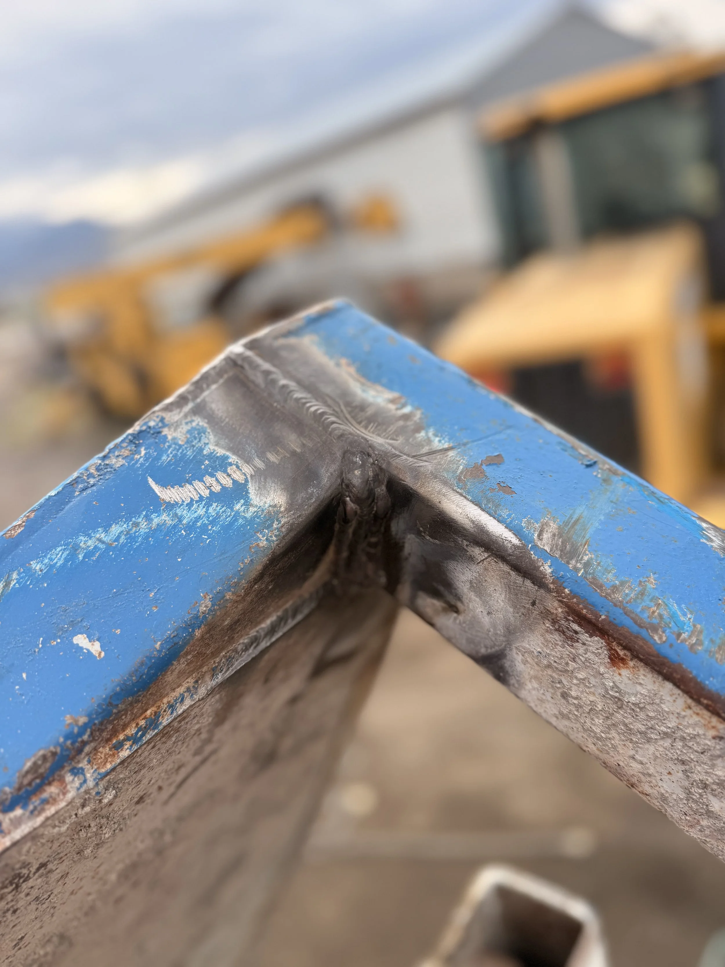 Close-up of a metal corner of a blue painted structure with visible welding, chipped paint, and rust, with construction equipment blurred in the background.