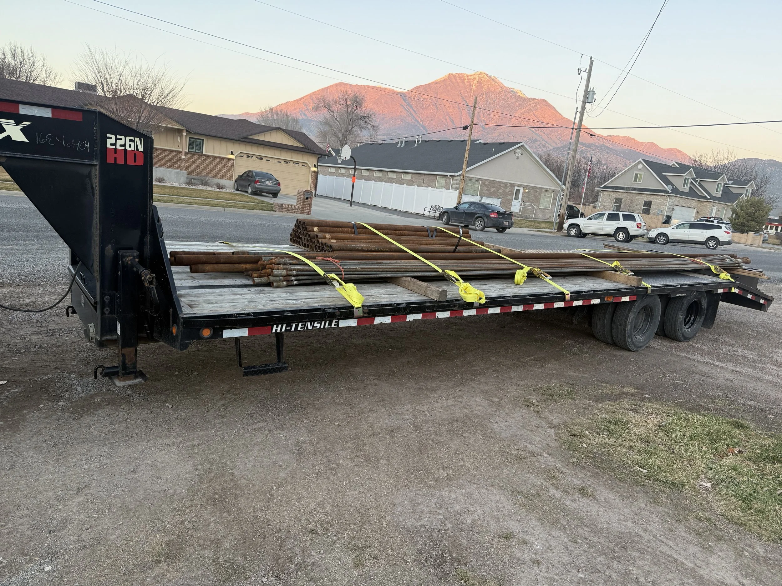 Flatbed trailer parked on gravel with bundles of metal pipes secured with yellow straps, residential houses, utility poles, and a mountain in the background at sunset.