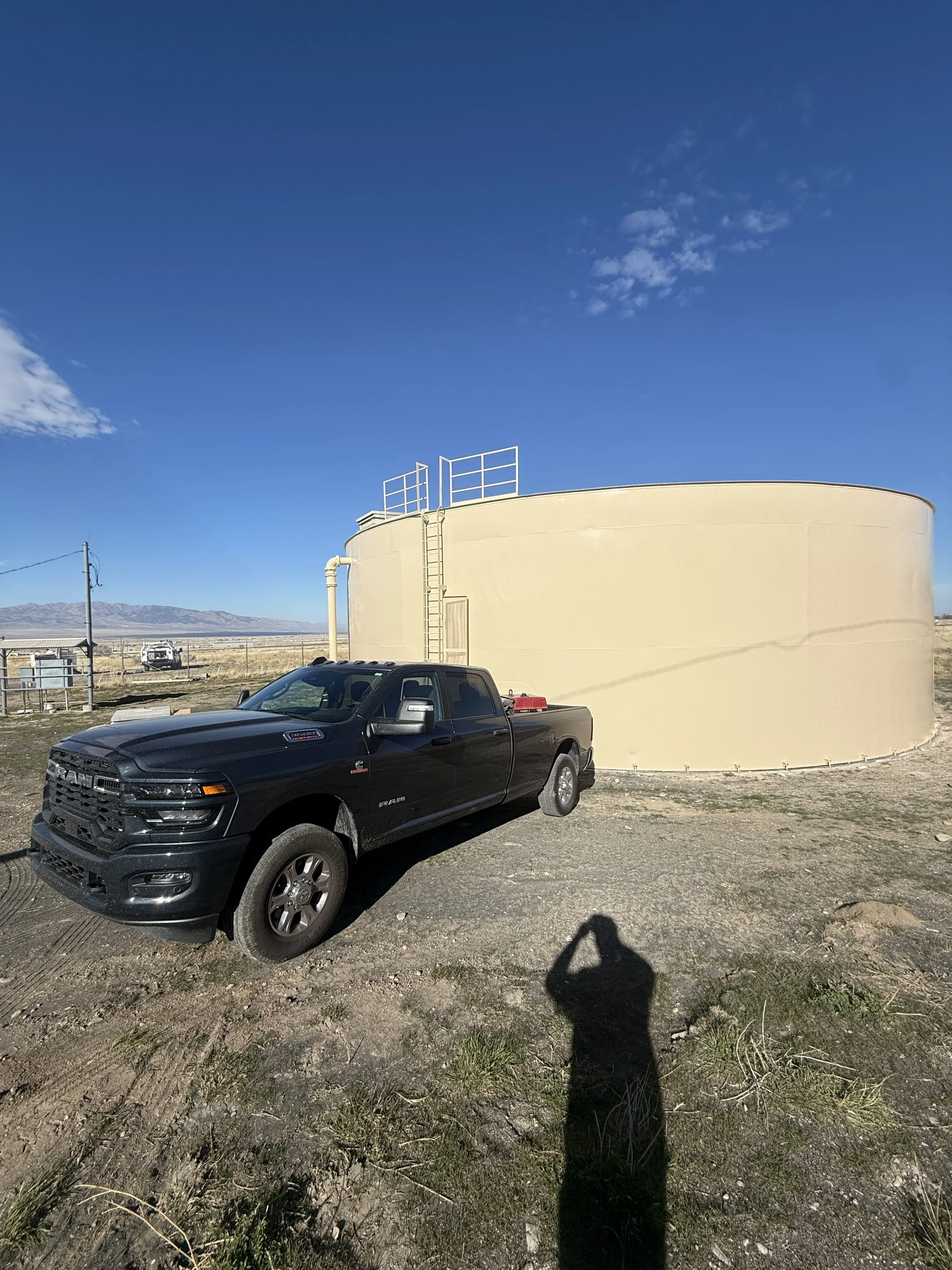 A black pickup truck parked in front of a large yellow tank in a rural area under a blue sky with some clouds.