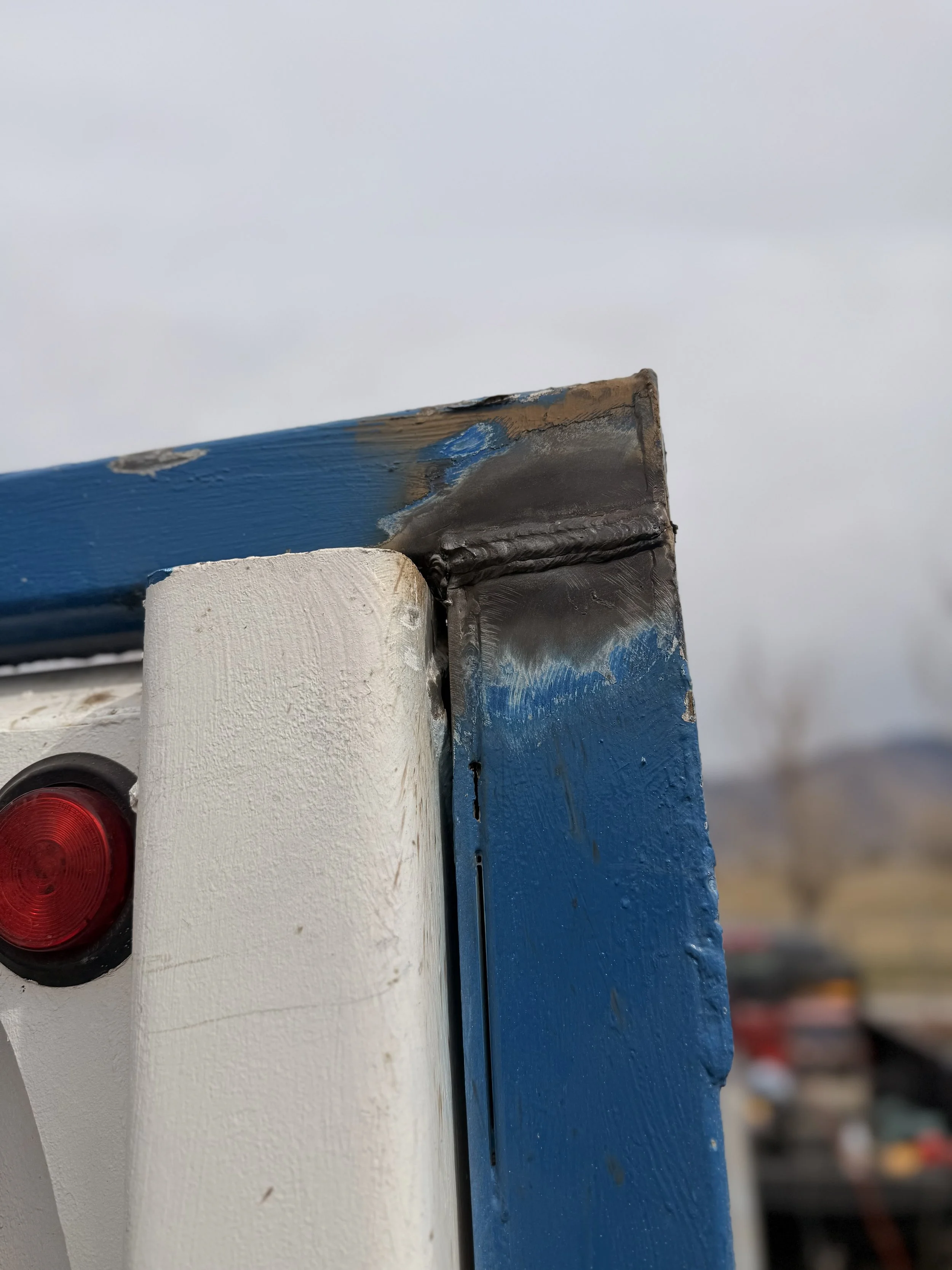 Close-up of the corner of a painted wooden structure, showing white, blue, and black paint with visible welding at the joint, set against an overcast sky.
