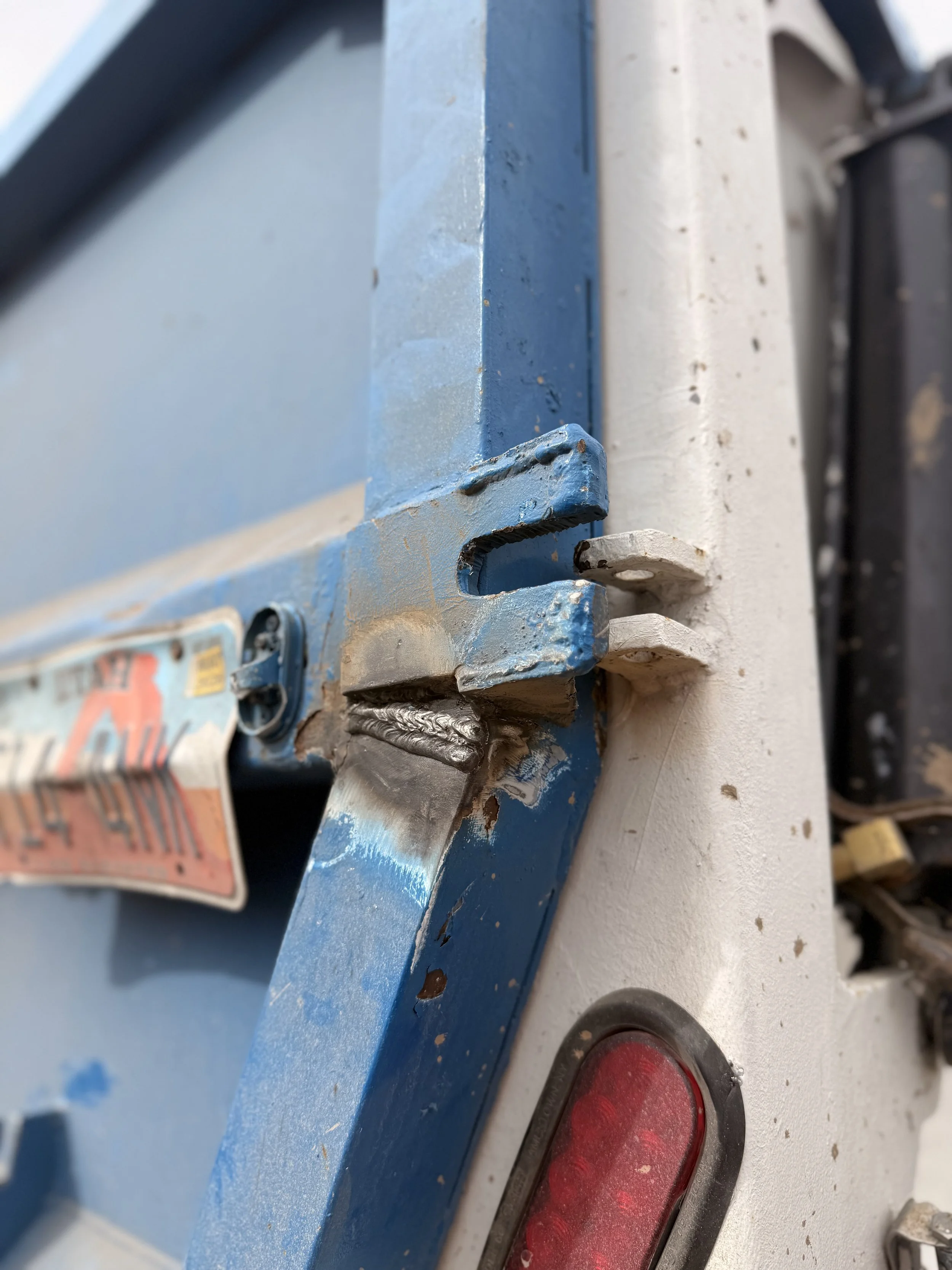 Close-up of a blue metal latch or hinge attached to a white surface, with a red taillight nearby, on a vehicle or machinery.