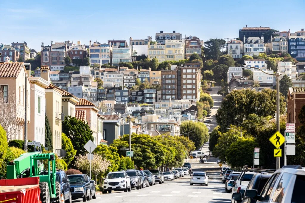 Street view of a hilly neighborhood with parked cars, residential buildings on hills, trees, and a pedestrian crossing sign.