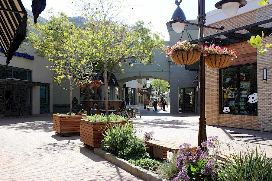 A shaded outdoor shopping plaza with trees in large wooden planters, hanging flower baskets, storefronts, and a brick building with a window display of toys and panda decorations.