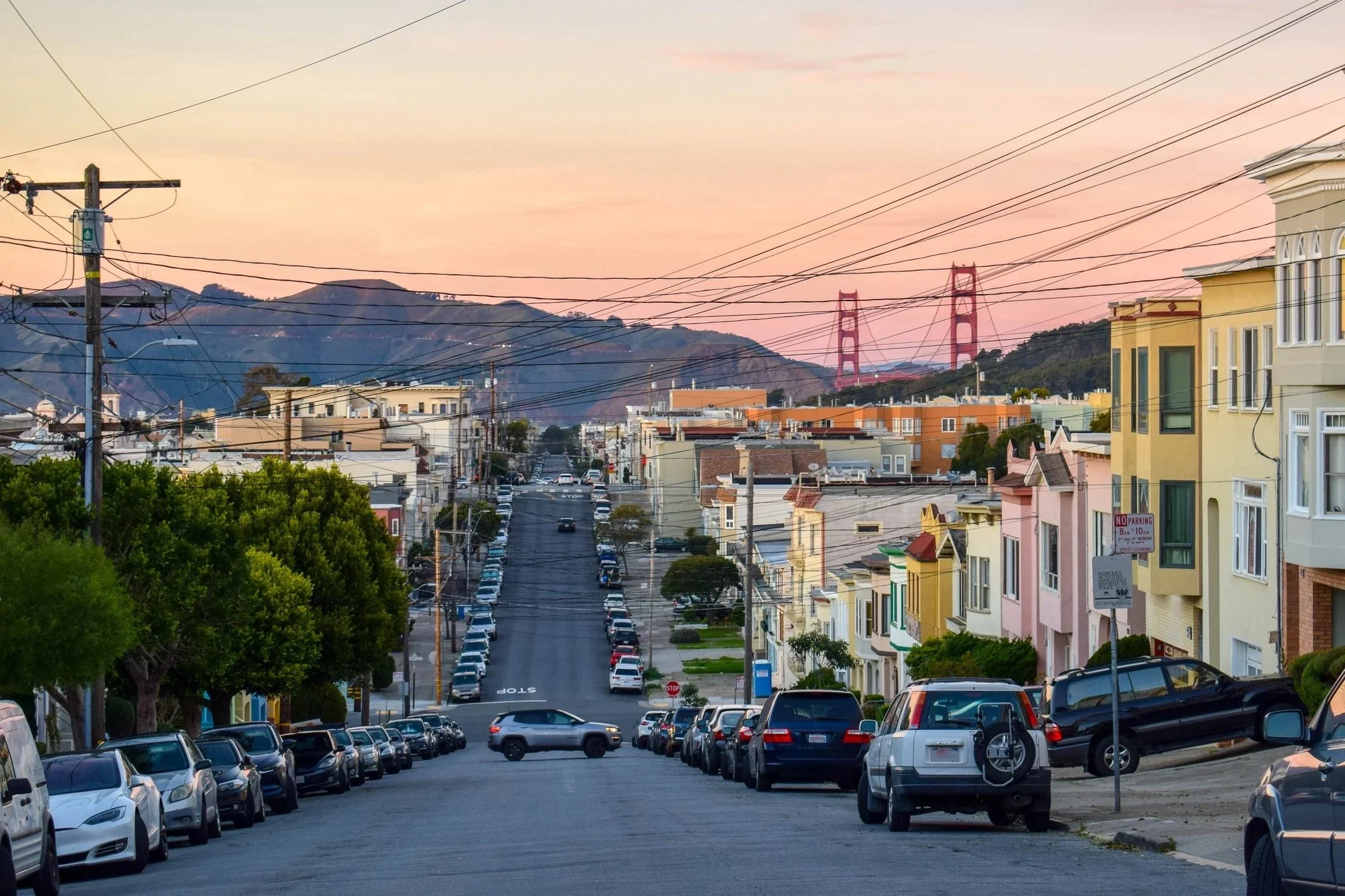 City street scene during sunset with parked cars along the street, multi-story residential buildings with colorful facades on the right, power lines crossing the sky, and a bridge visible in the background over hilly terrain.