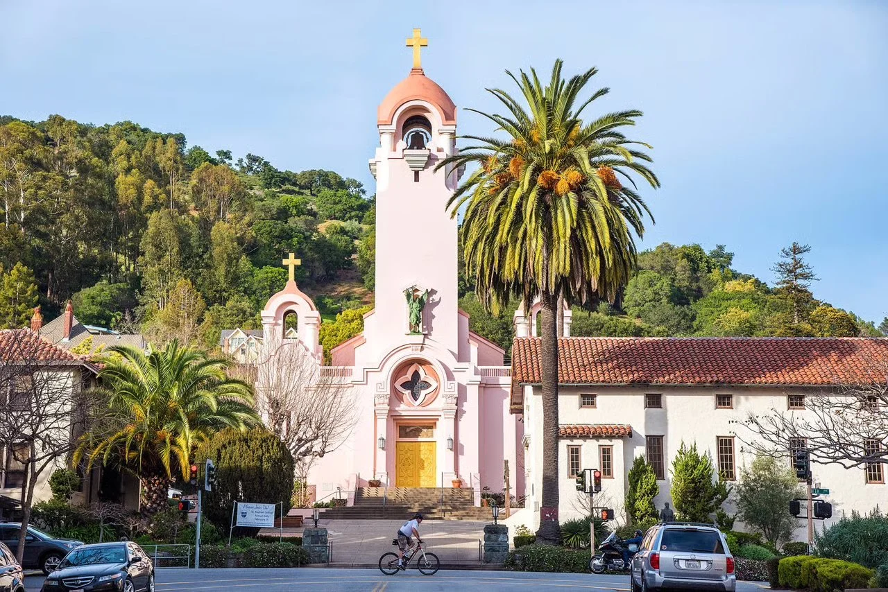 A small white church with a tall bell tower topped with a cross, featuring pastel pink accents and surrounded by palm trees, with cars and a bicyclist in the foreground.