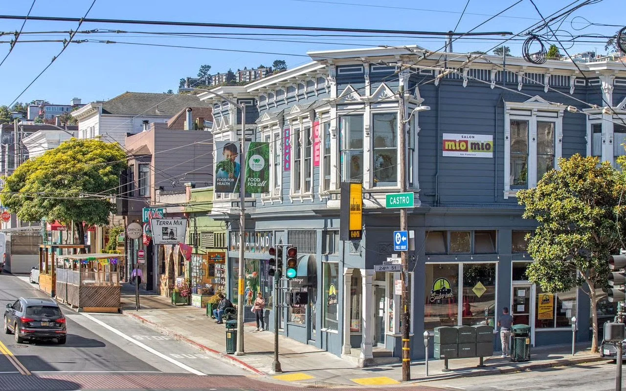 Street scene at the corner of Castro Street with a blue building housing a salon called Mio Mio, traffic lights, pedestrians, cars, and storefronts, including a shop named Terra Mía.