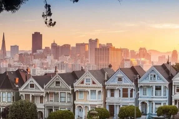 Beautiful row of Victorian-style houses with a city skyline in the background during sunset.