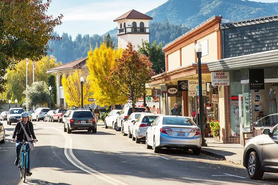 A street scene in a small town with parked cars, pedestrians, and a cyclist, surrounded by colorful trees and buildings with mountain scenery in the background.