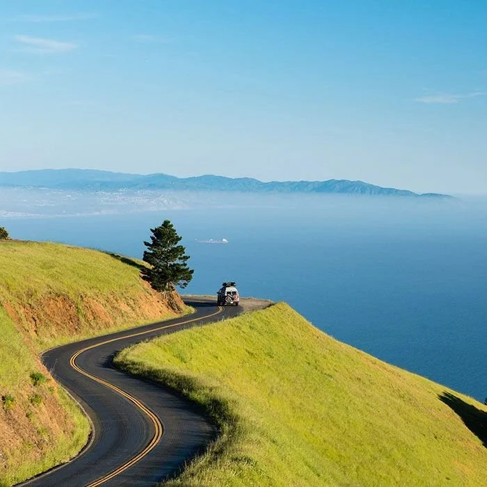 A winding road along green hills overlooking the ocean with a ship and distant mountains in the background.
