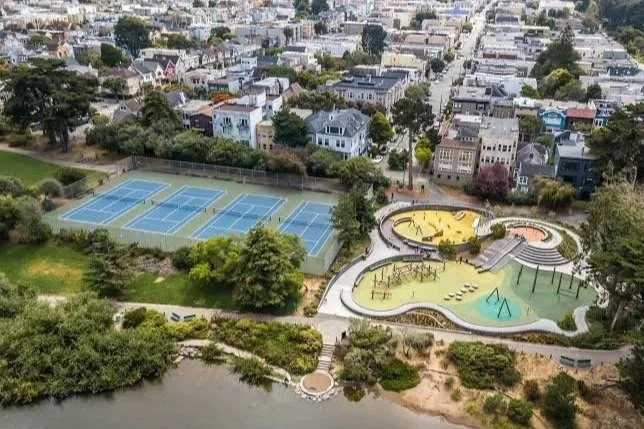 Aerial view of a park with tennis courts, playgrounds, and a small body of water surrounded by trees and residential buildings.