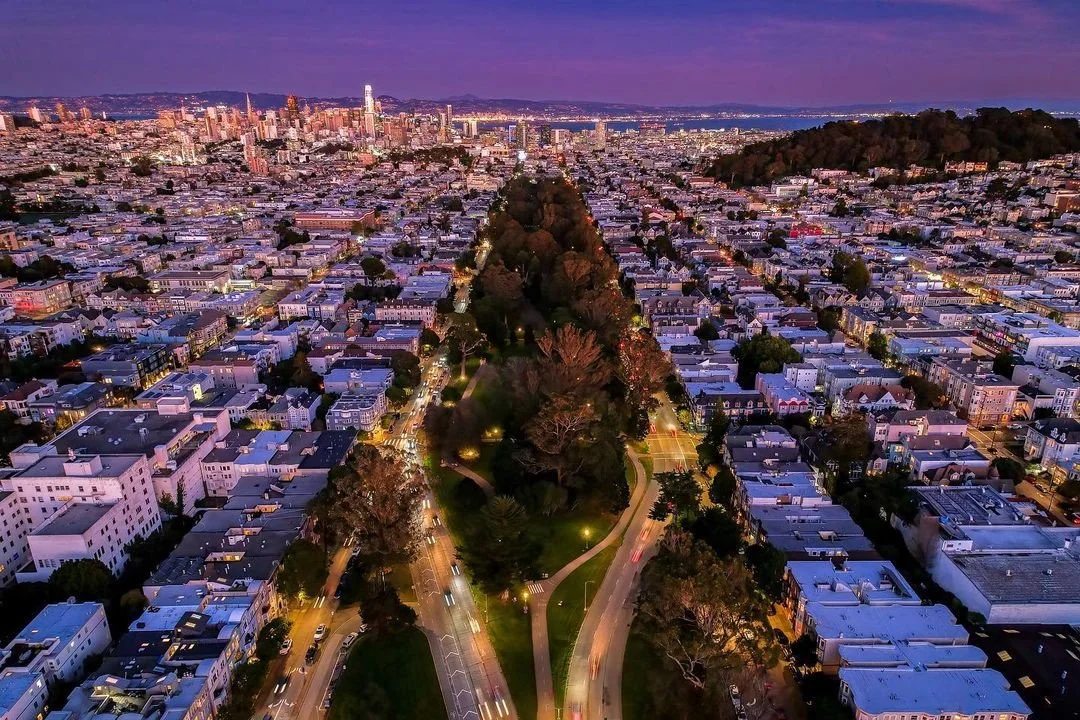 Aerial view of a city at dusk with a long, tree-lined park in the center, surrounded by tightly packed residential buildings, with a skyline featuring tall skyscrapers in the background.