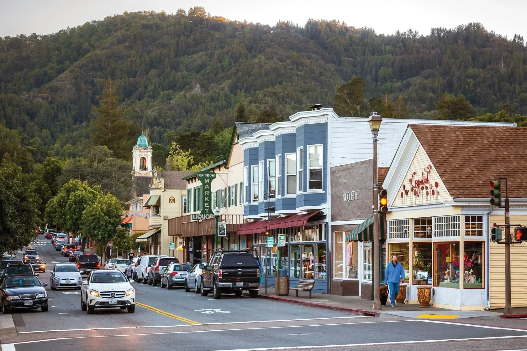 A small town street with colorful buildings, parked cars, and a traffic light. In the background, there are green, forested hills and a church steeple with a blue roof. The street appears busy with some pedestrians.
