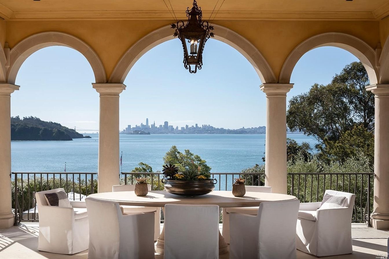Outdoor patio with a round dining table, six chairs, decorative pots, overlooking a water view with city skyline in the distance.