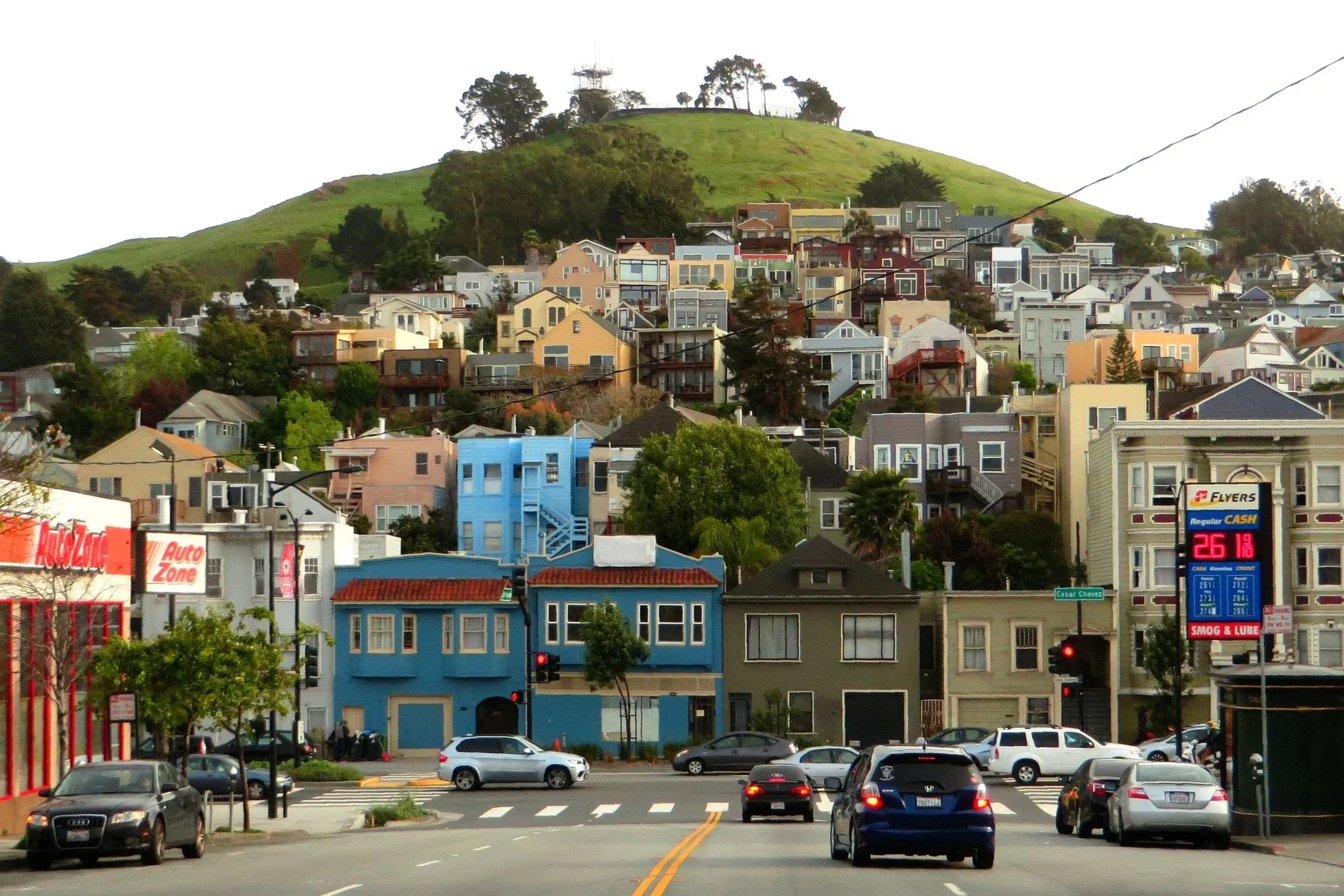 City street with cars, colorful houses on a hill, and a green hill with trees and an antenna on top in the background.