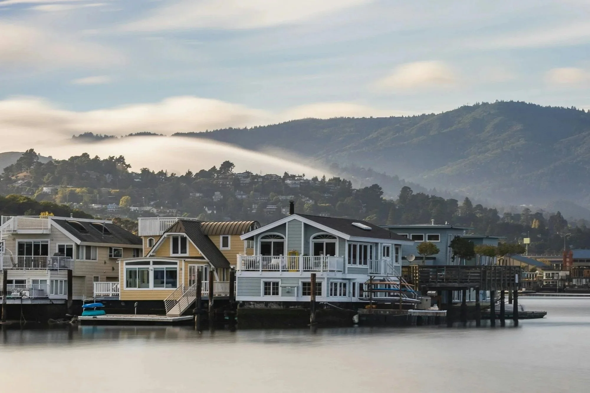 Colorful houses with large windows and balconies along a waterfront, with boats docked nearby and a misty mountain backdrop under a partly cloudy sky.
