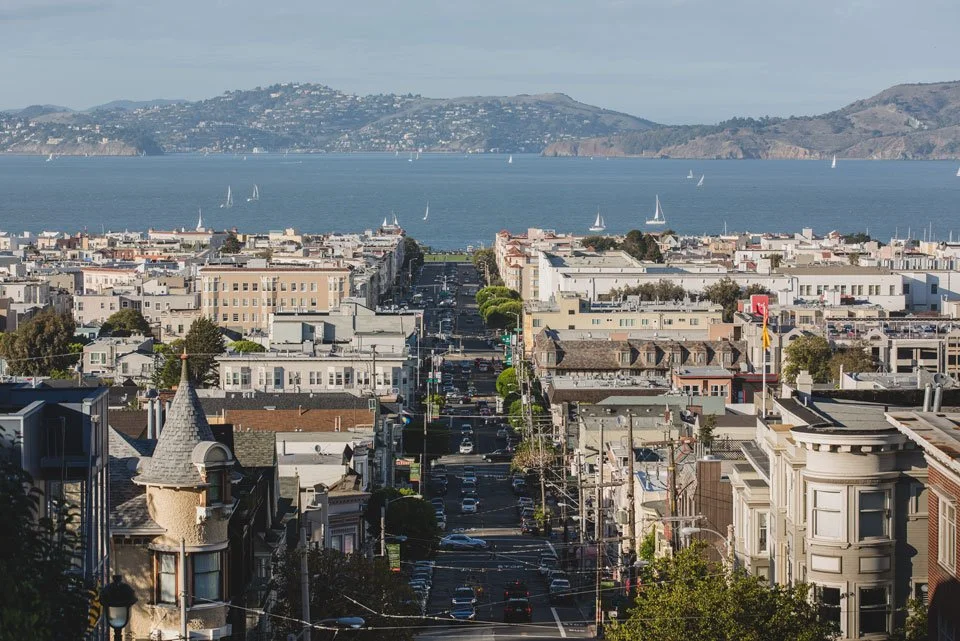 A cityscape view overlooking a street in San Francisco, with the bay and sailboats in the background.