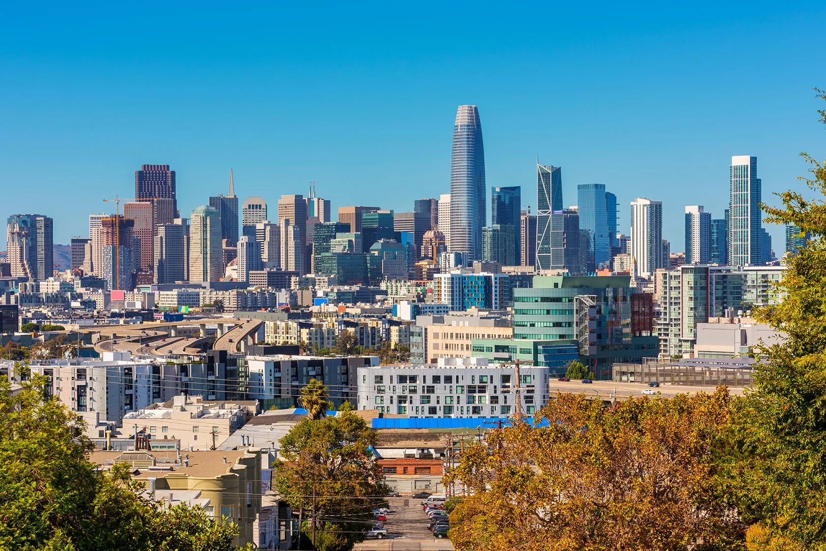 A cityscape featuring tall skyscrapers and modern buildings under a clear blue sky, with some trees in the foreground.