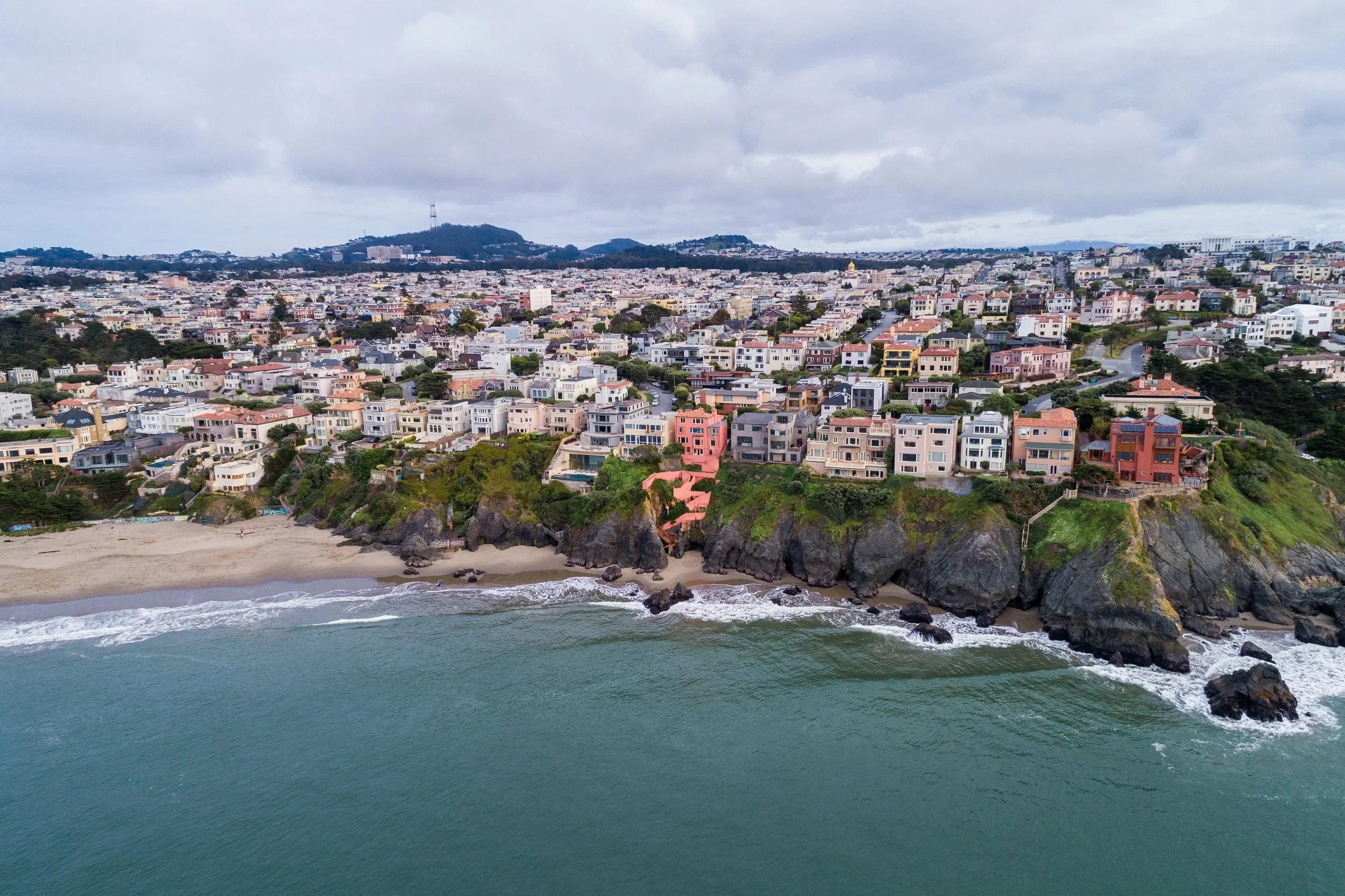A panoramic view of a city with numerous houses and buildings on a hilly terrain near the ocean, with waves crashing onto rocks along the shoreline. The sky is cloudy.