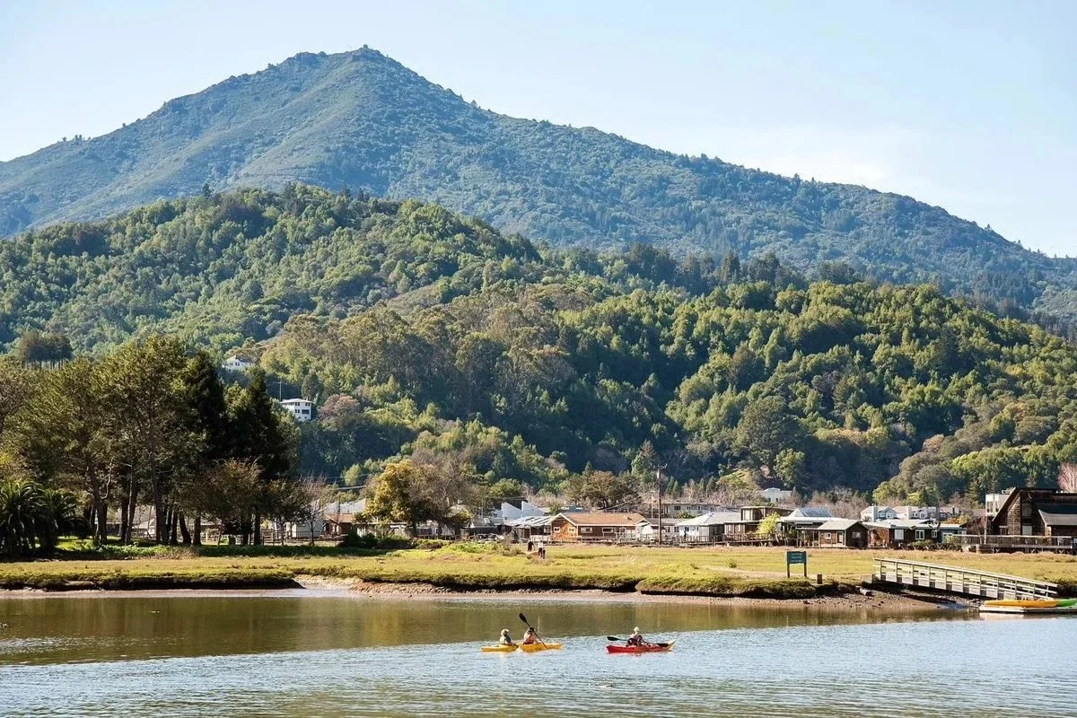 Scenic view of a mountain with lush greenery, houses at the base, a calm river in the foreground with people kayaking, and a clear sky.