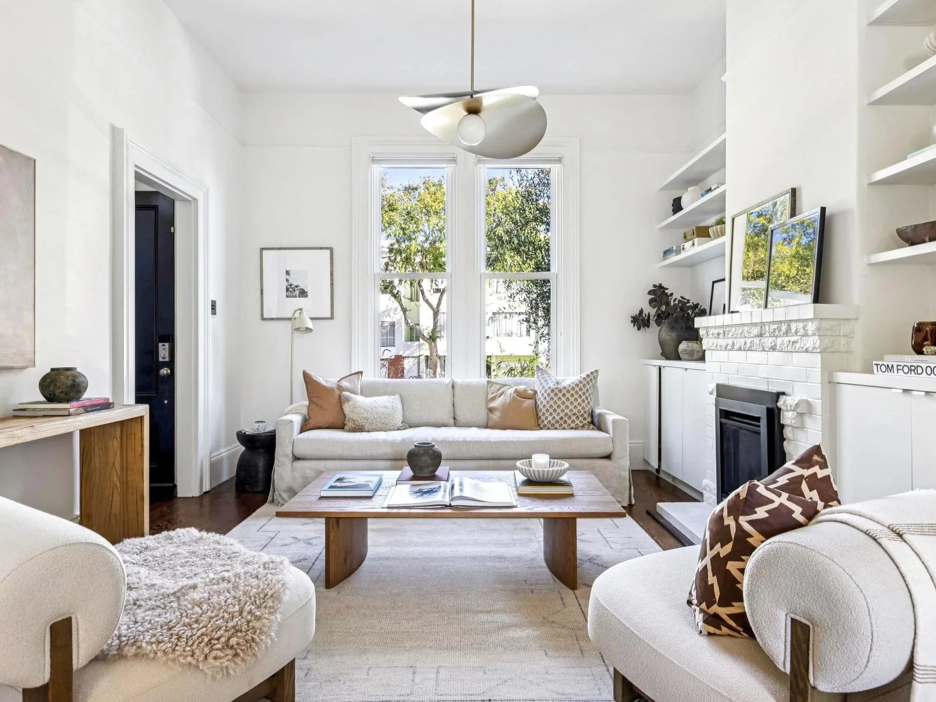 Bright living room with white walls, large windows showing trees outside, white sofa with pillows, wooden coffee table with books and decor, white fireplace with a mirror on mantel, and modern ceiling light fixture.