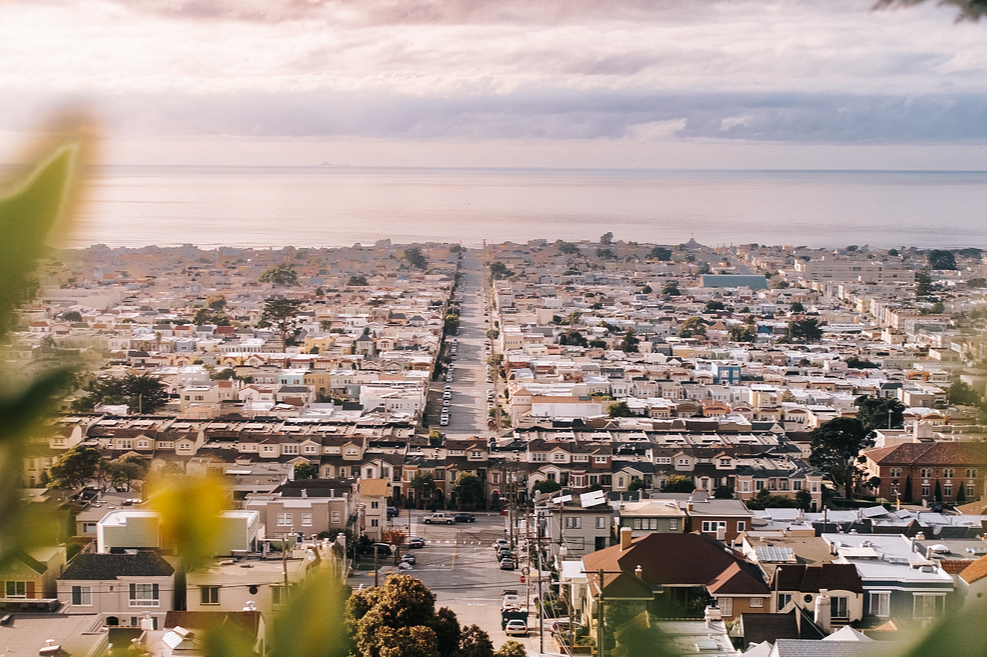 Aerial view of a densely populated coastal city with grid-like streets, houses, and buildings, overlooking the ocean in the distance under a cloudy sky.