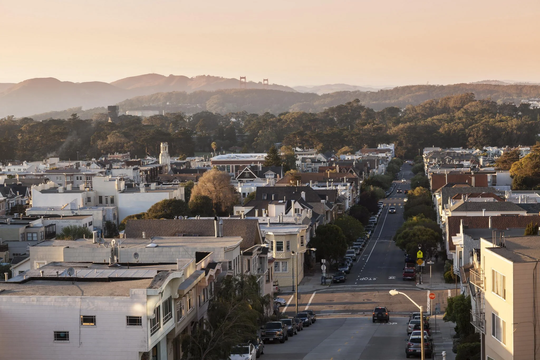 Sunset over a residential neighborhood with houses, trees, and a distant view of hills and a bridge in the background.