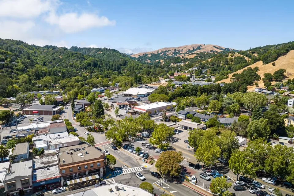 Aerial view of a small town surrounded by green hills and mountains, with a mix of commercial and residential buildings, parking lots, and tree-lined streets.