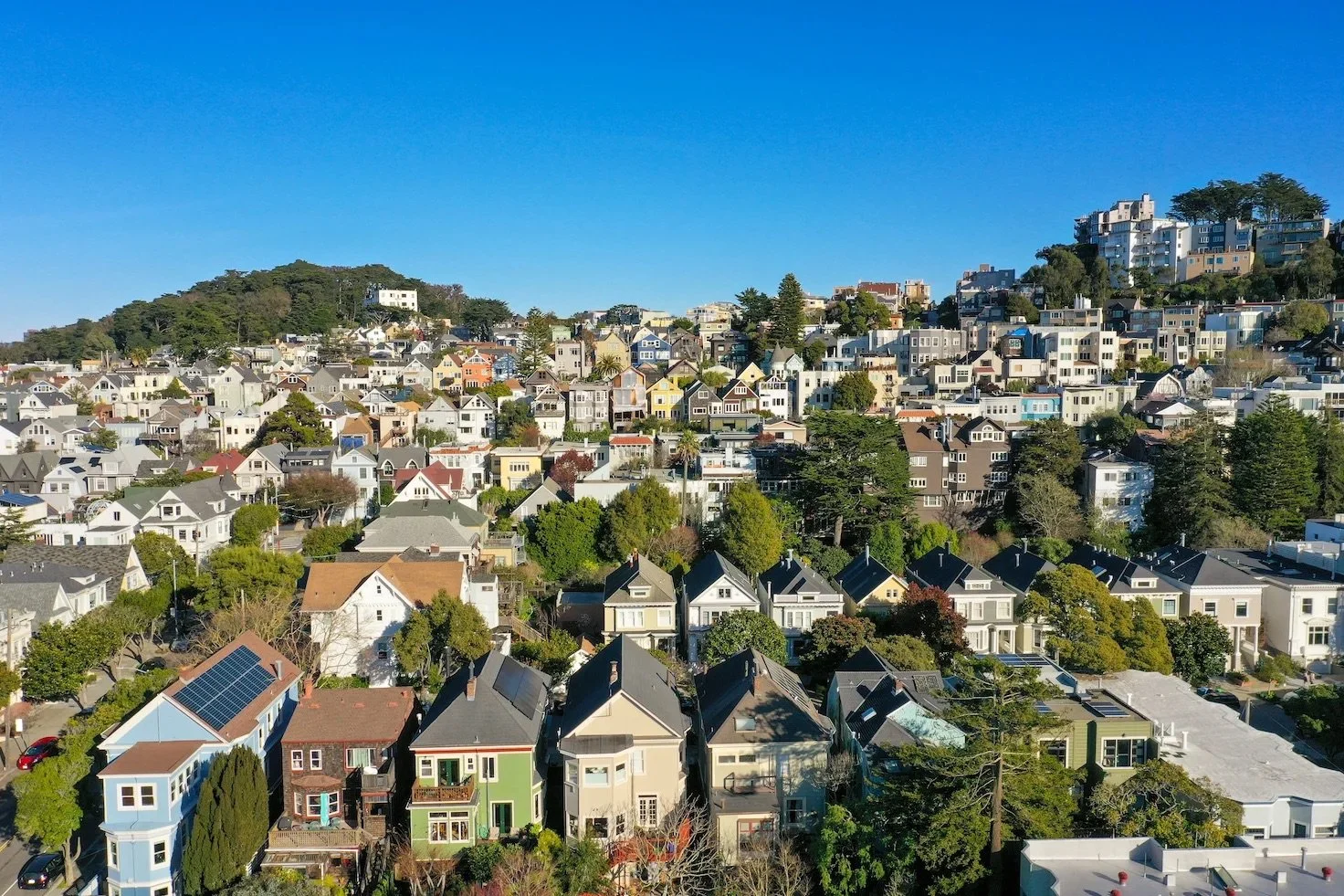 Aerial view of a densely packed hillside residential neighborhood with a variety of colorful houses and green trees, under a clear blue sky.