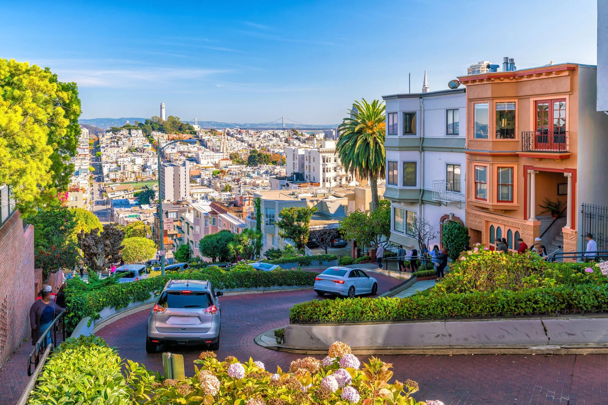 View of San Francisco cityscape with colorful row houses, palm trees, cars parked along a winding brick road, and the bay bridge in the background under a clear blue sky.