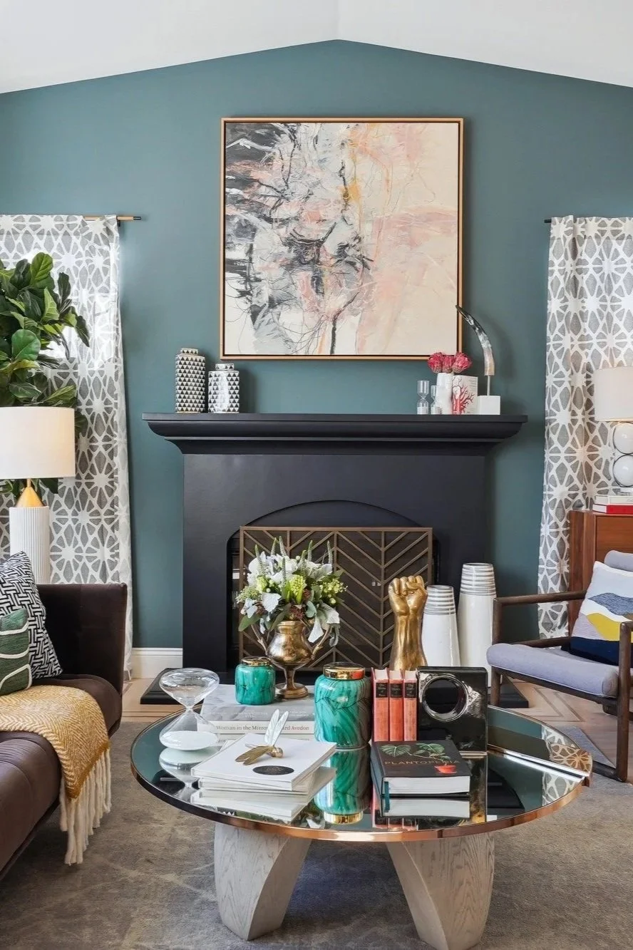 Living room with teal wall, black fireplace, abstract artwork, patterned curtains, green plant, white lamps, decorative vases, flowers, books, and modern decor.