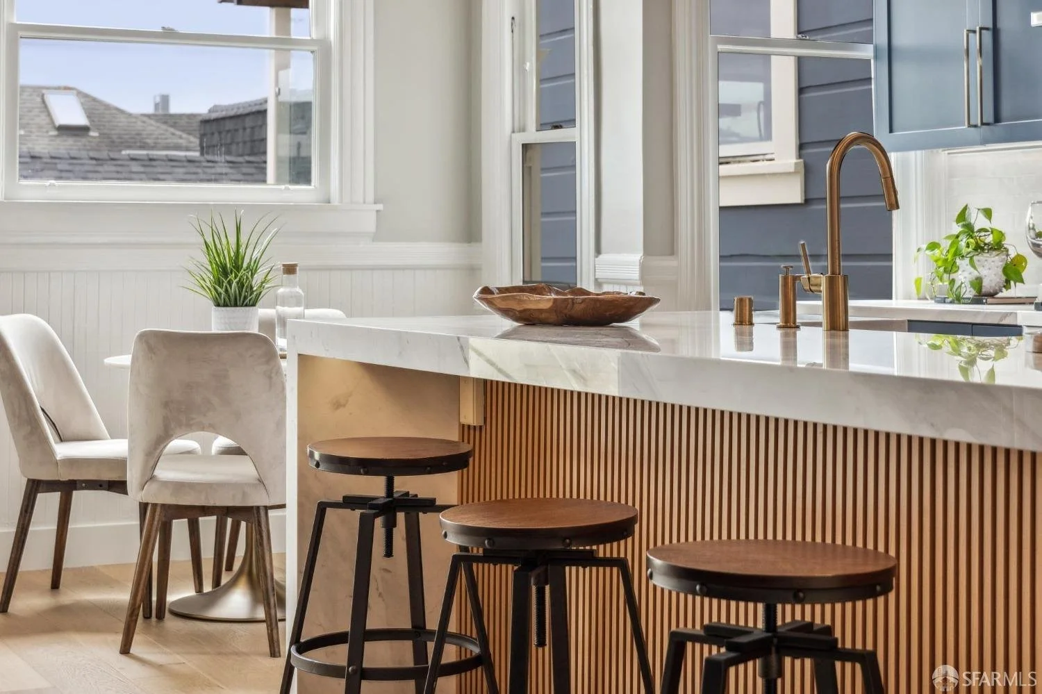 Modern kitchen with white marble island and copper fixtures, beige and dark wood bar stools, white chairs, green plants near window, and blue cabinets.
