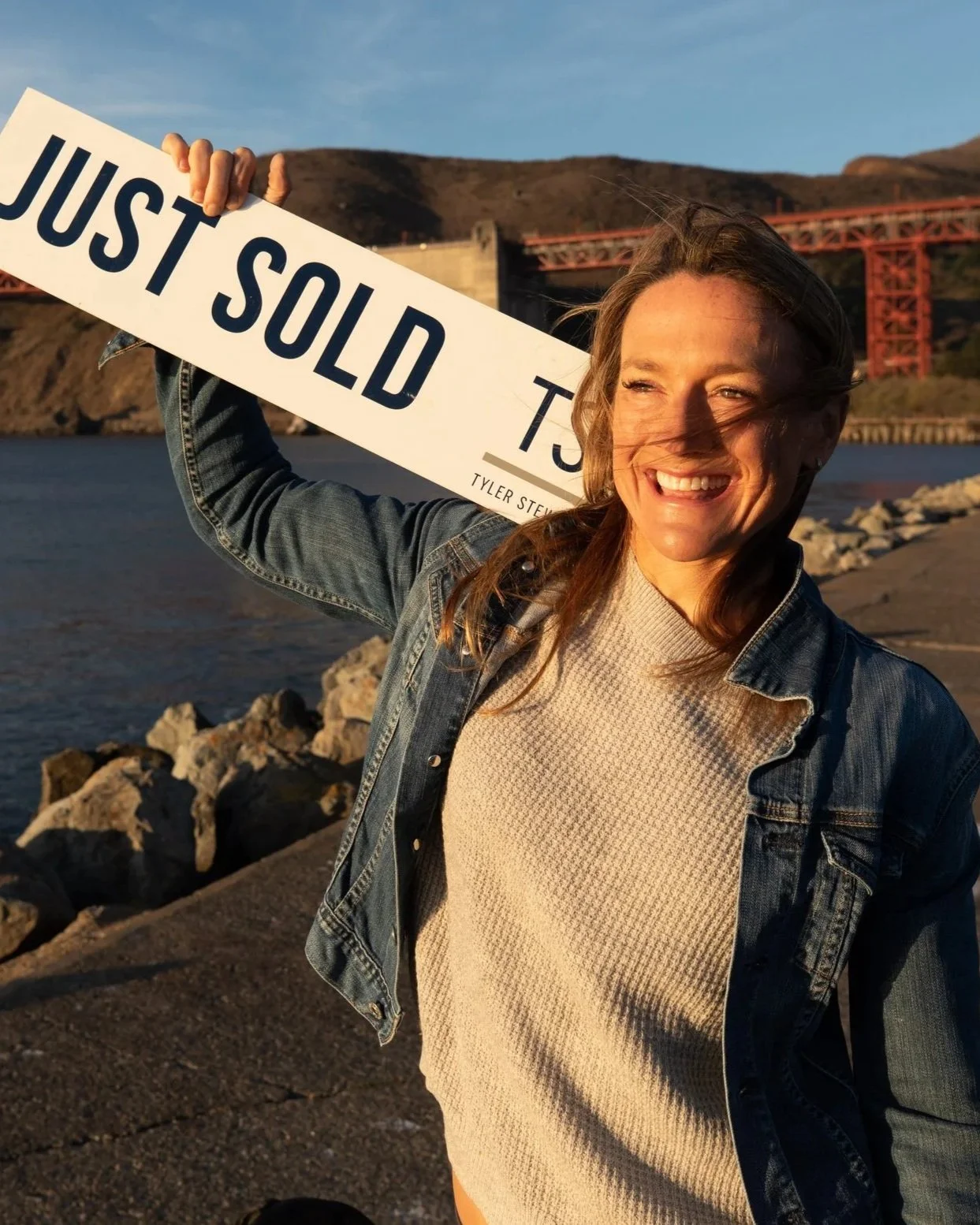 Woman smiling outdoors holding a 'Just Sold' sign with a bridge and hilly landscape in the background during sunset.