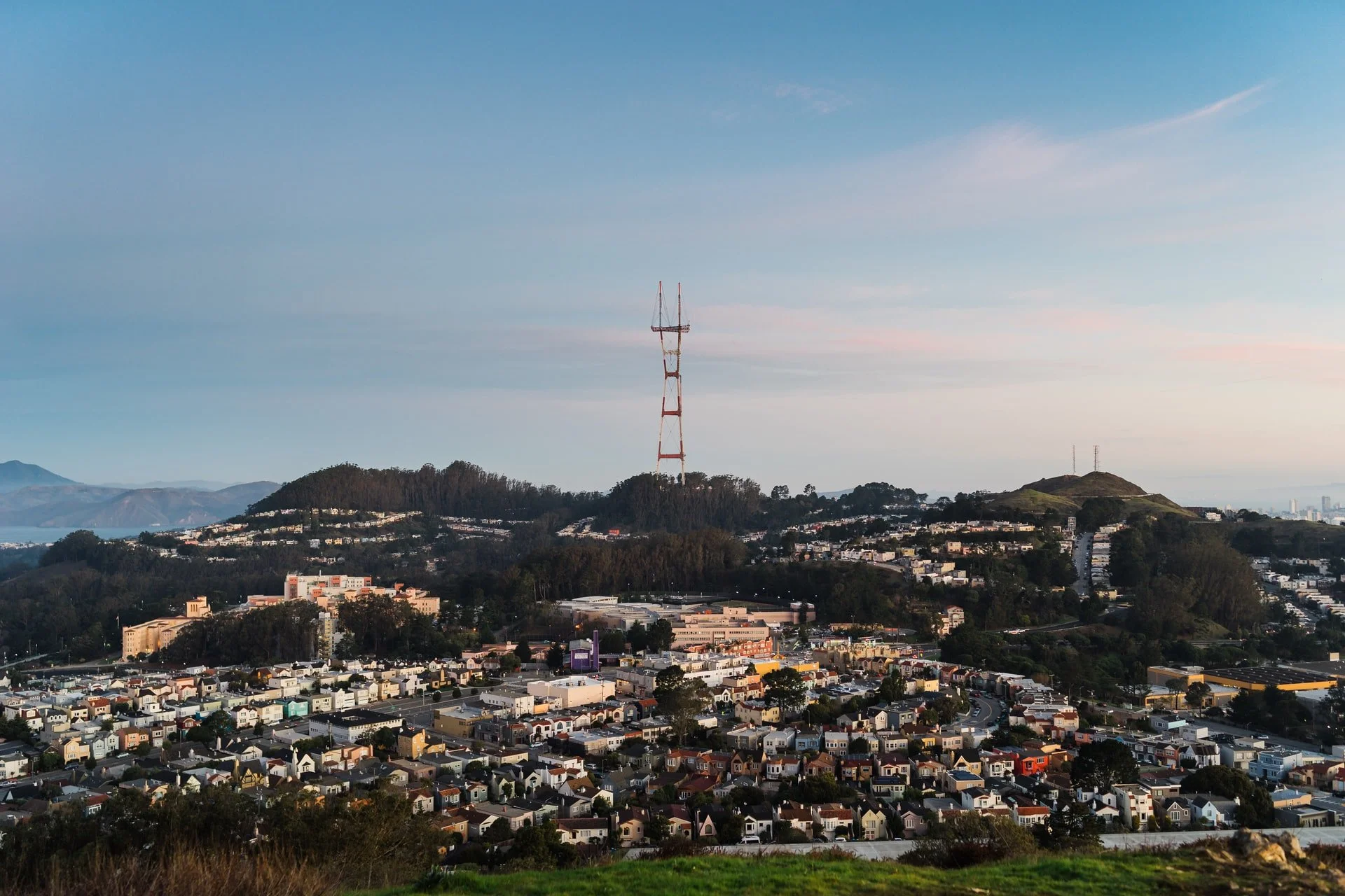 A view of a cityscape with a hilltop and a tall radio tower, residential buildings, and a skyline in the distance under a partly cloudy sky.