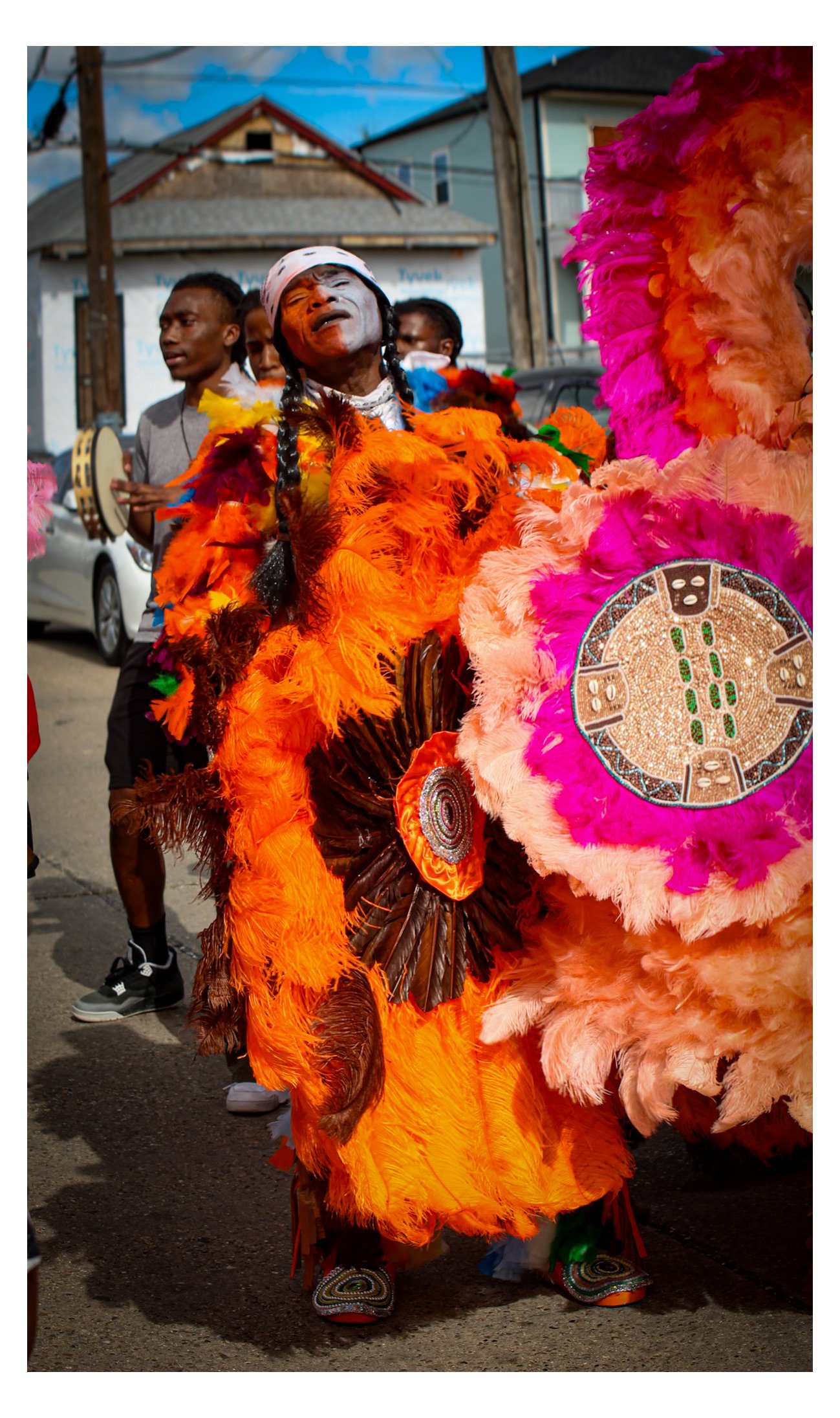 A person dressed in vibrant orange and pink feathered costume, wearing a headdress with beadwork, during a cultural street parade, with other participants and houses in the background.