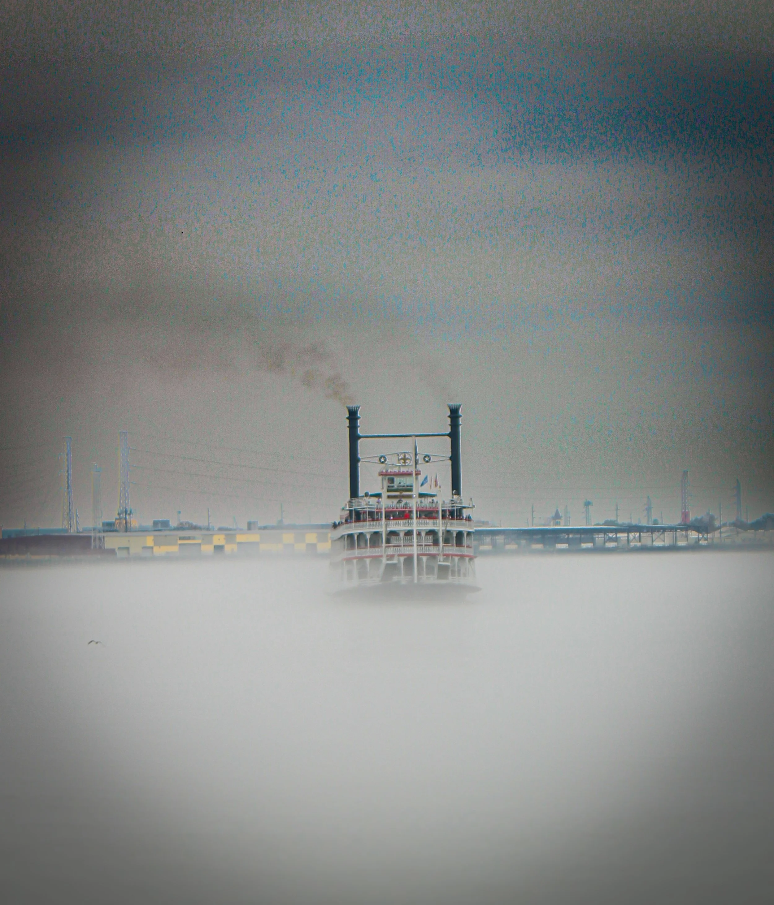 A large industrial facility with two tall smokestacks emitting smoke, situated near a body of water with a bridge in the background.