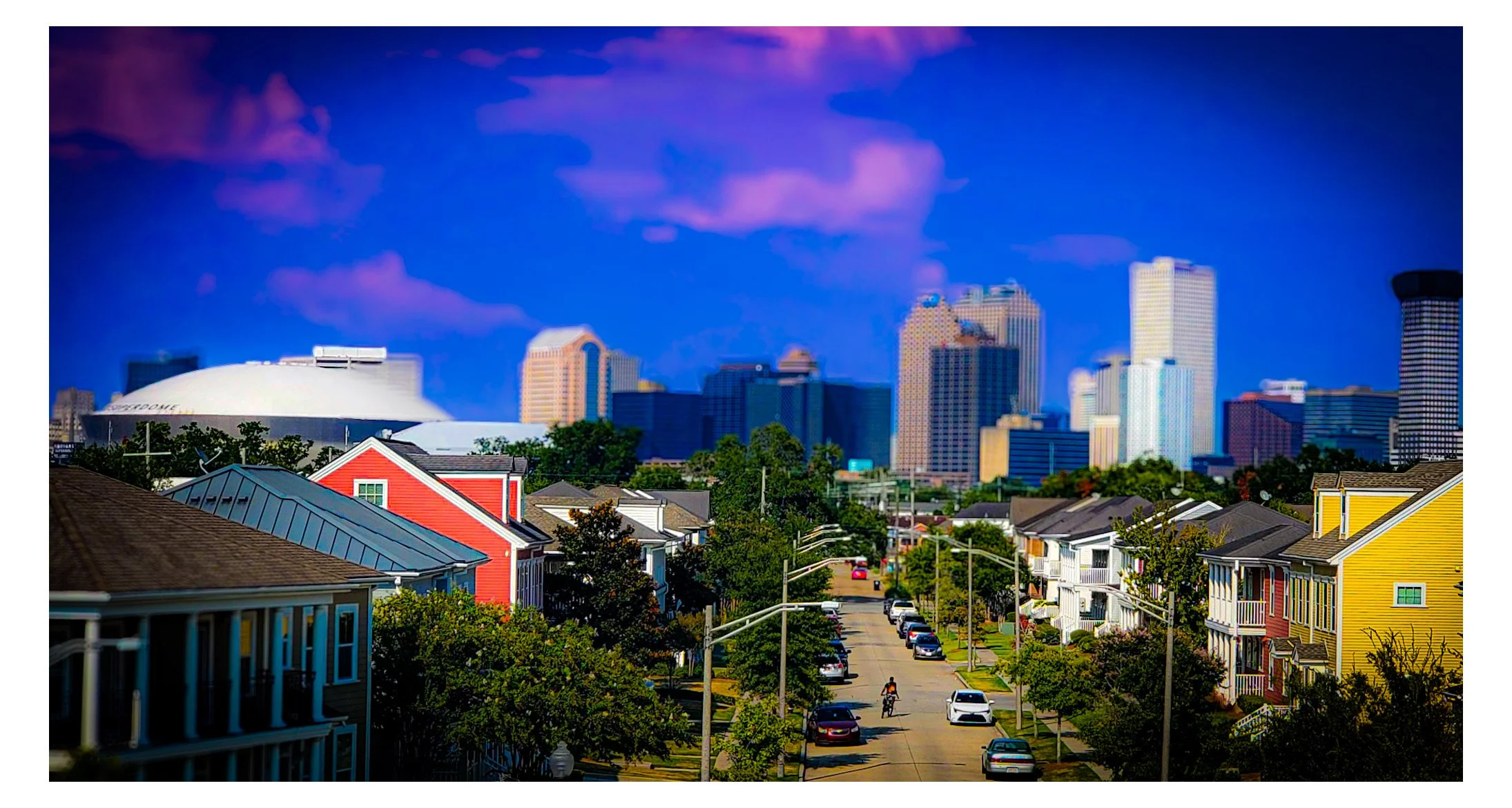 Colorful neighborhood with houses and a street in foreground, and a city skyline with tall buildings and a domed sports arena in background, under a partly cloudy blue sky.