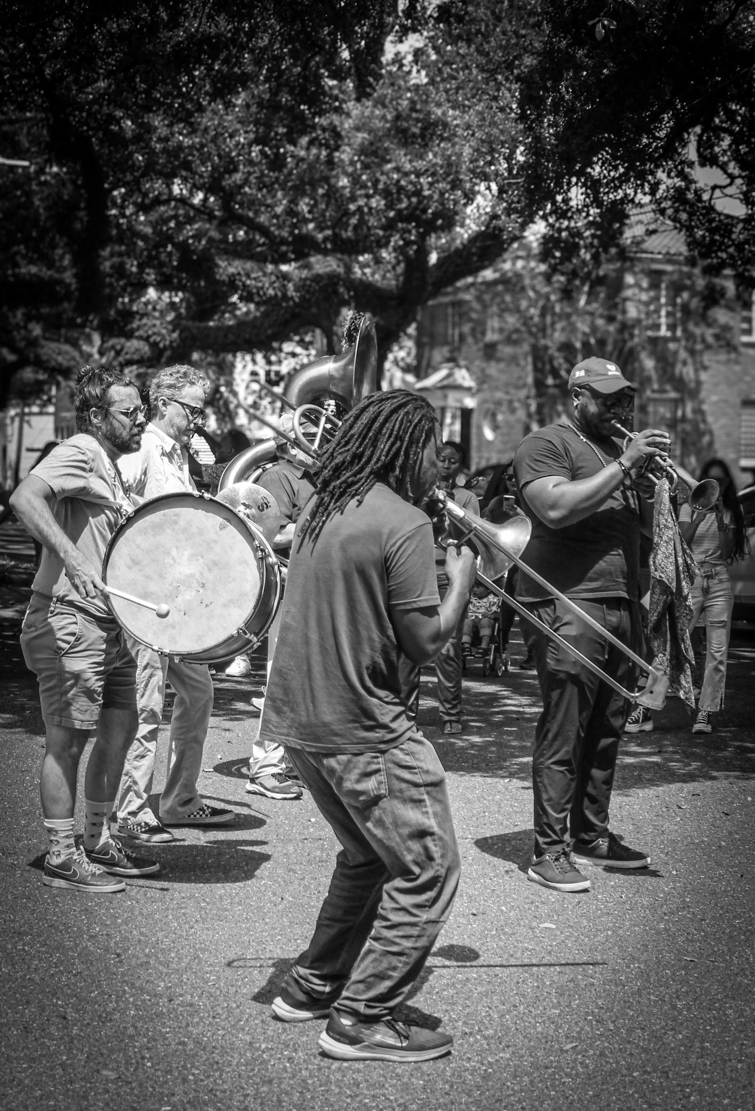 Black and white photo of a group of street musicians playing instruments outdoors, including a large drum and saxophonists, with trees and buildings in the background.