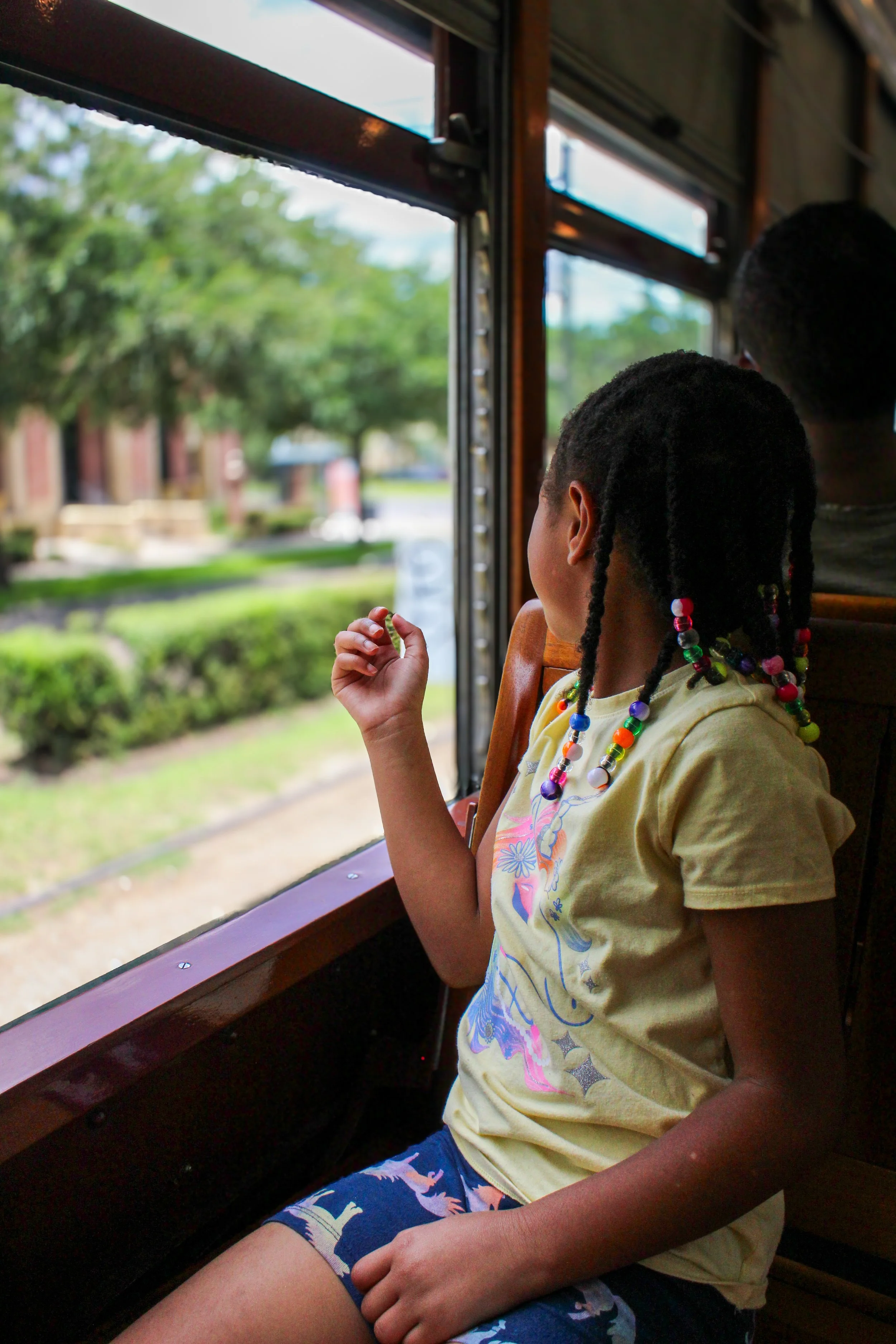 A young girl with braided hair and colorful beads is looking out a window on a train or bus. She is wearing a yellow t-shirt with a colorful design and shorts with dinosaur patterns. Outside, there are trees and buildings visible.