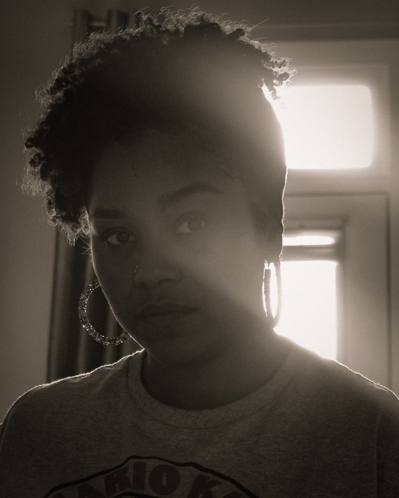 A woman with short, curly hair and hoop earrings standing in front of a bright window, with sunlight creating a silhouette effect.