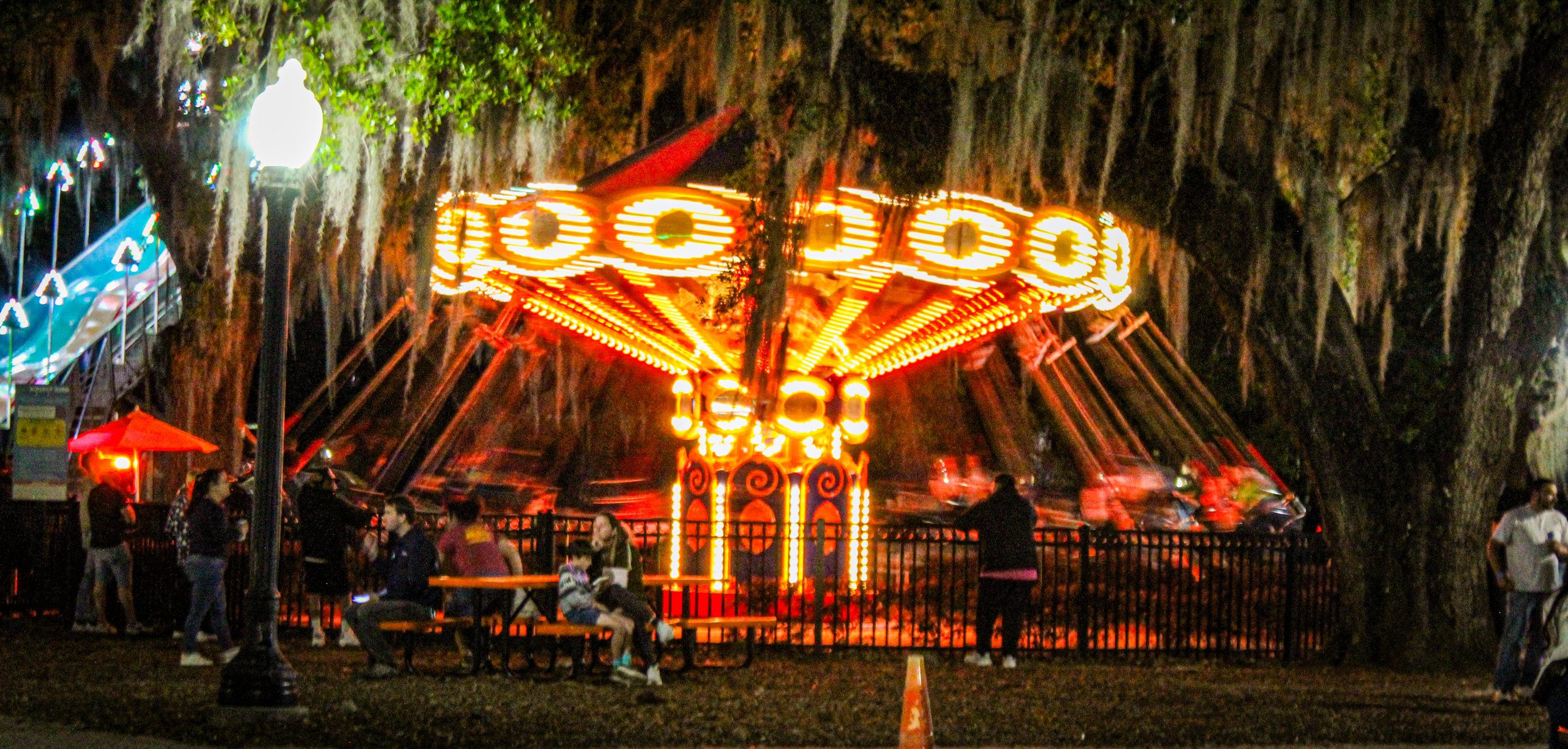 Night scene of a brightly lit carnival ride with orange and yellow lights, surrounded by people sitting on benches and walking nearby, with trees in the background.