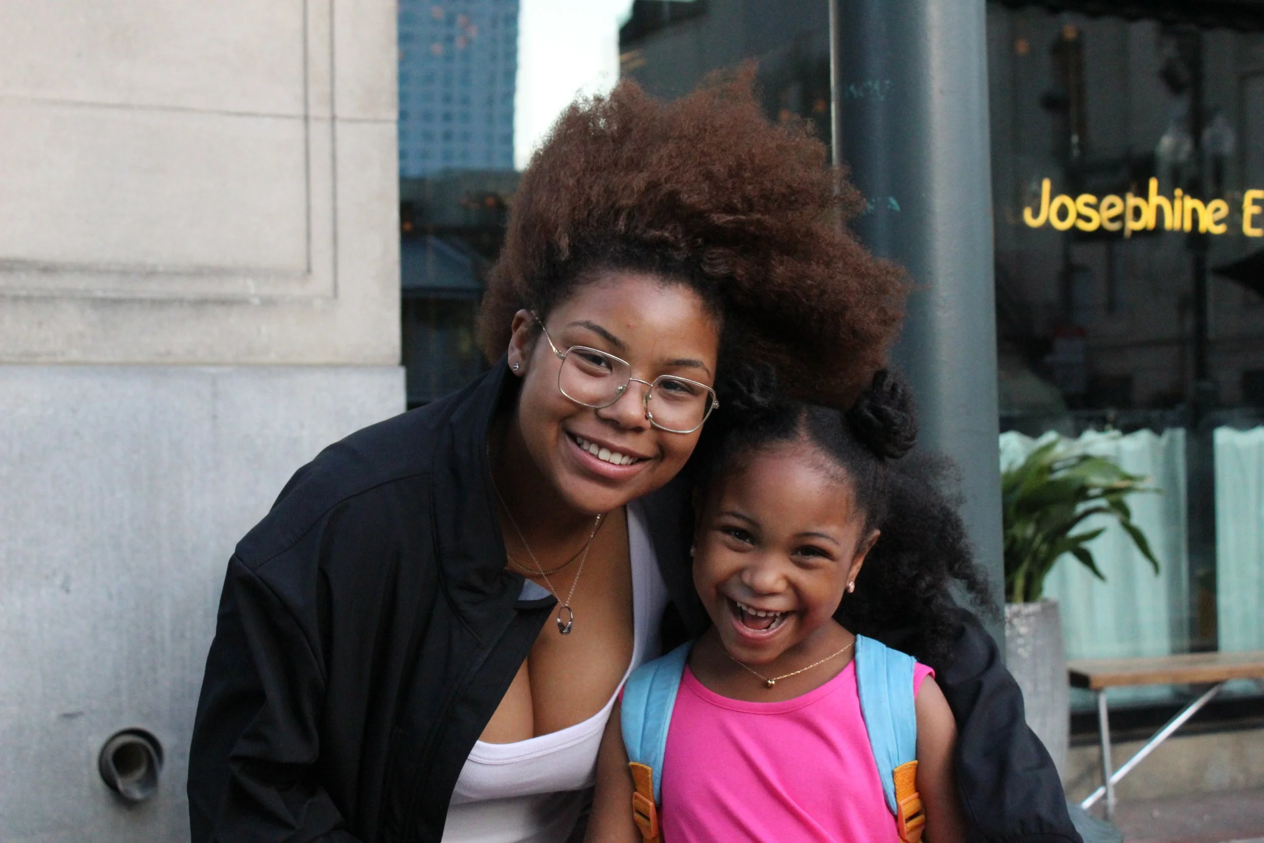 A smiling woman with curly reddish-brown hair and glasses, hugging a young girl with dark curly hair and a pink shirt with a blue backpack, standing outdoors in an urban setting.