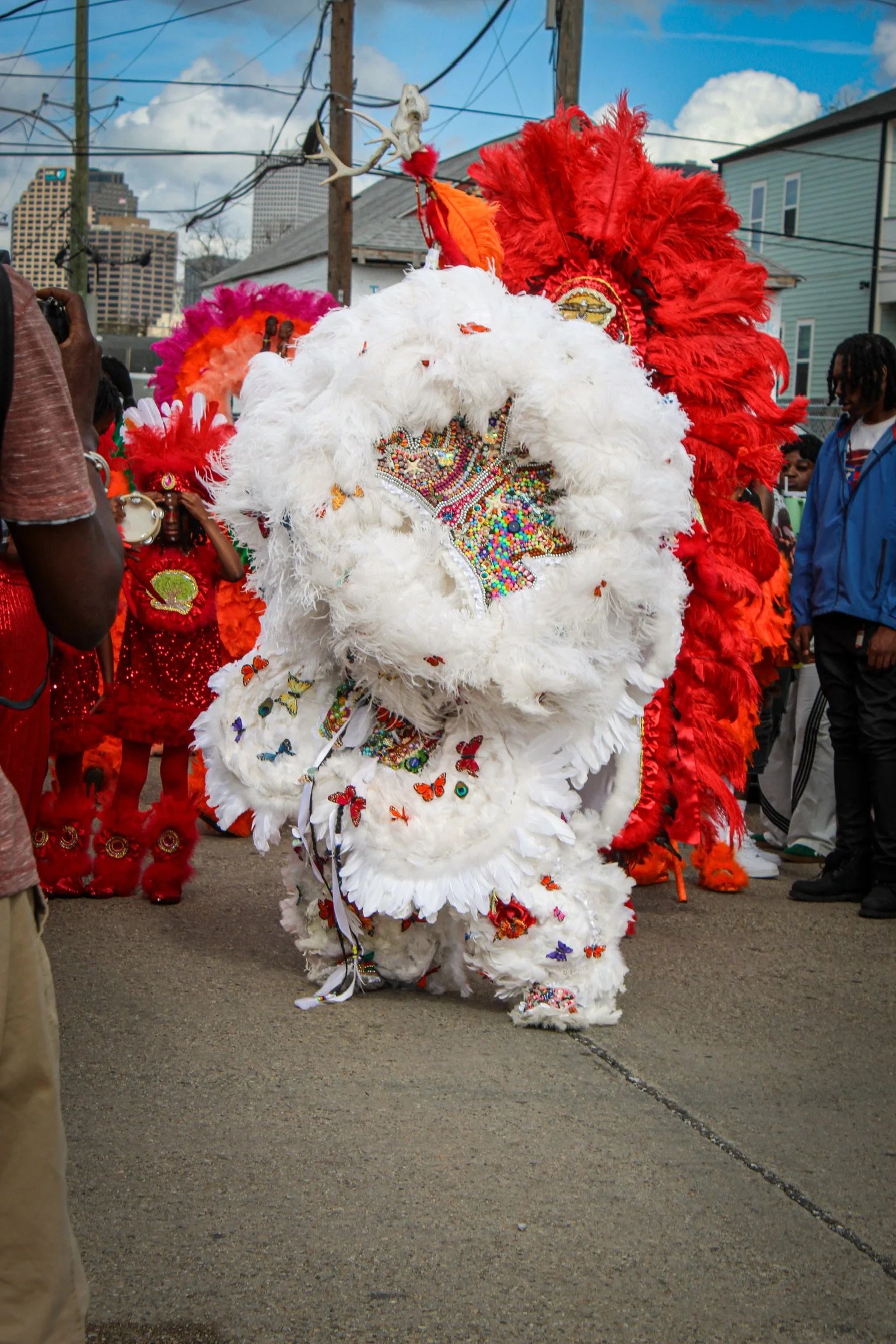 Person dressed in a traditional colorful costume with feathers, beads, and butterflies, participating in a cultural celebration parade, surrounded by other participants and spectators in an urban setting.