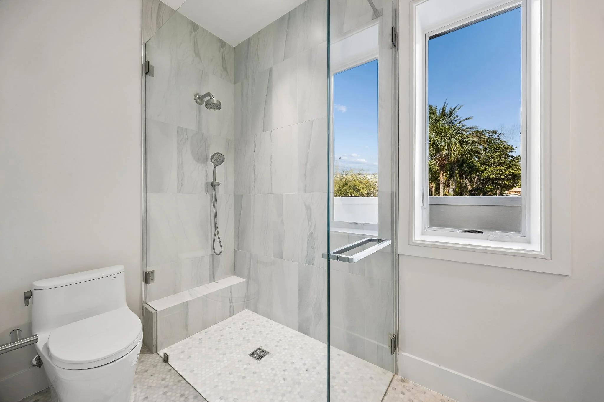 Modern bathroom with a glass-enclosed shower, toilet, and a large window showing a view of trees and a blue sky.