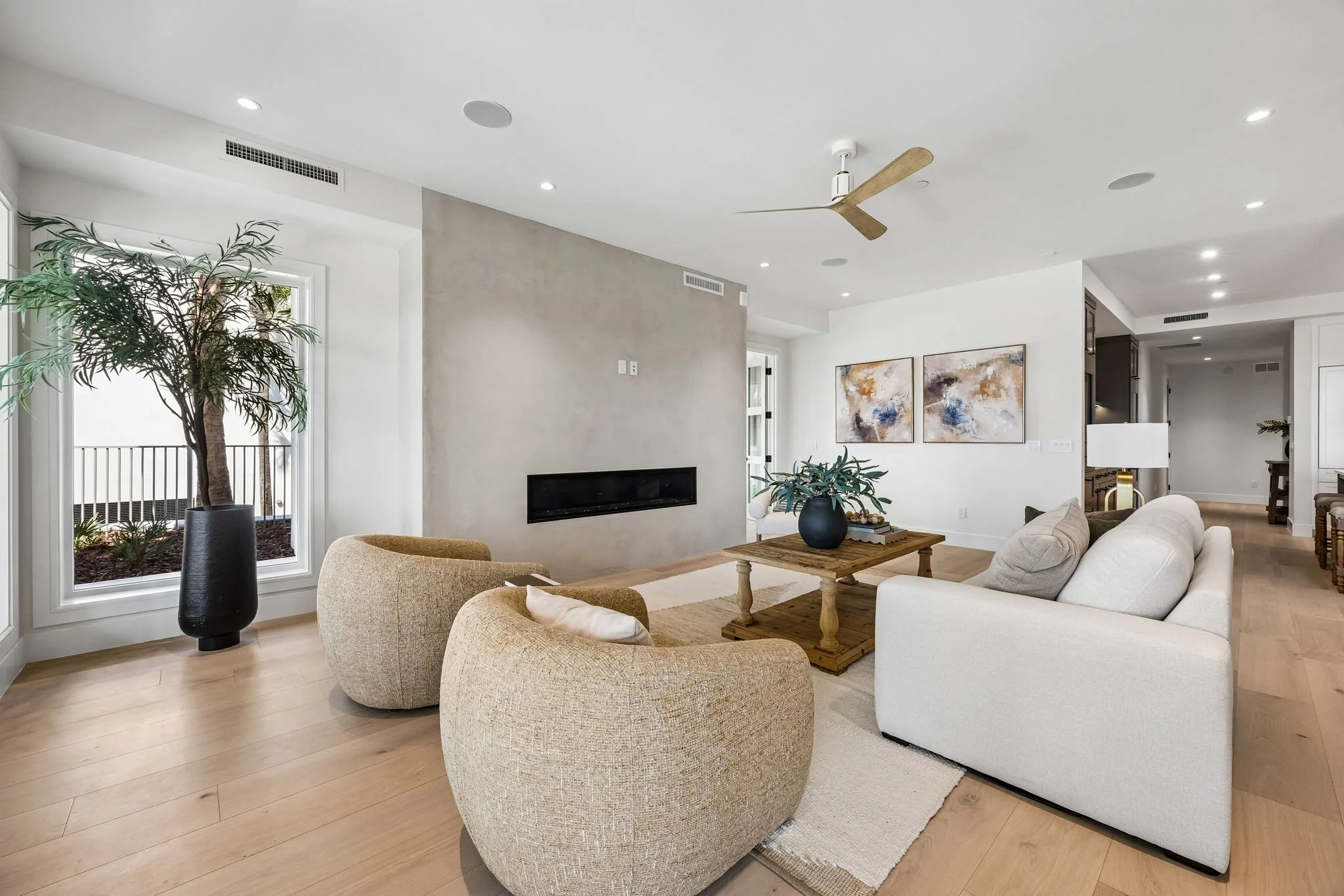 Modern living room with a sofa, two armchairs, a wooden coffee table with a plant, a fireplace, and wall-mounted abstract art, natural light from a window with a potted plant, and a ceiling fan.