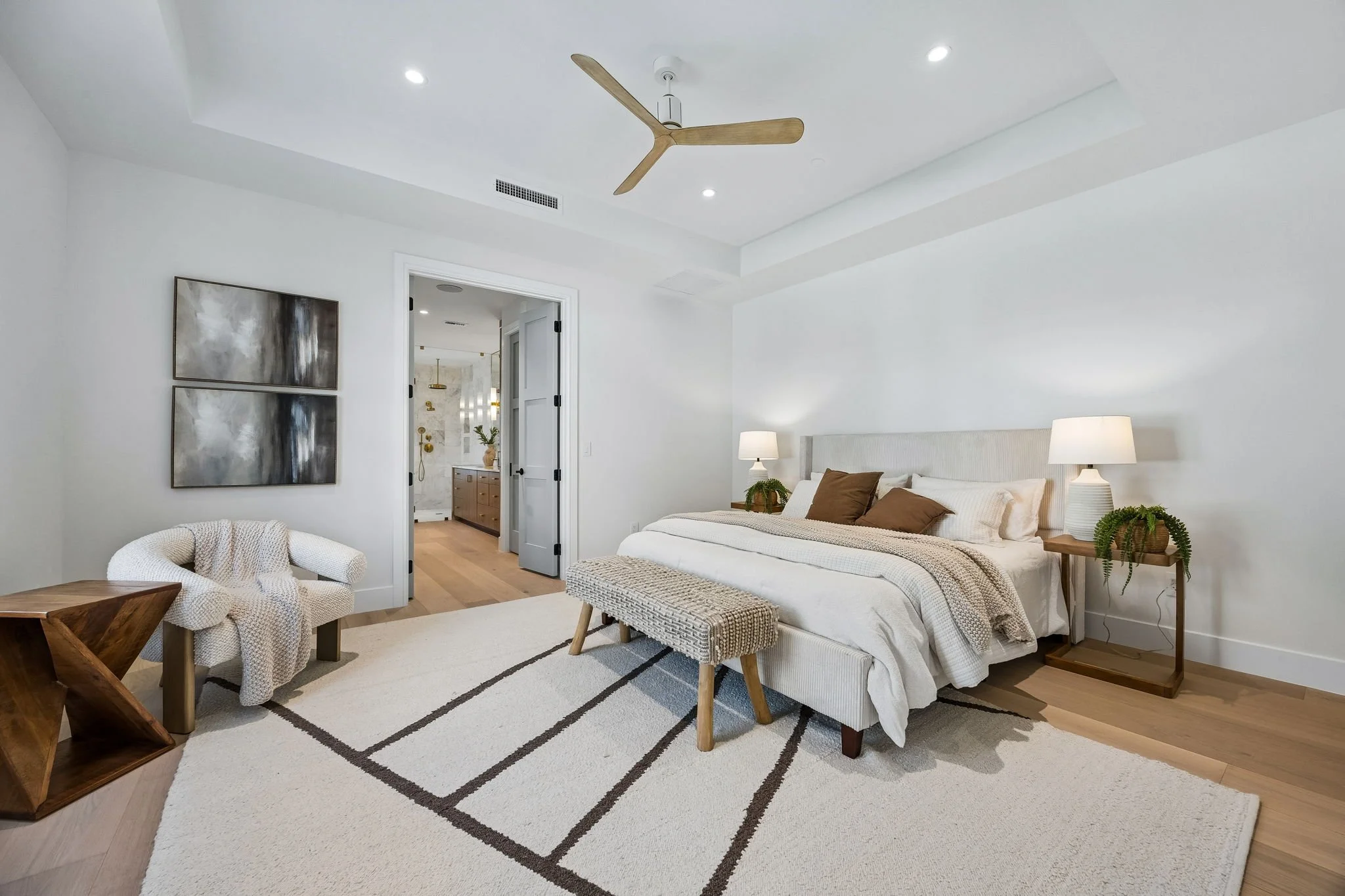 Modern bedroom with white walls, a large bed, bedside lamps, artwork, a bench, a cozy armchair, and an open doorway leading to a bathroom.